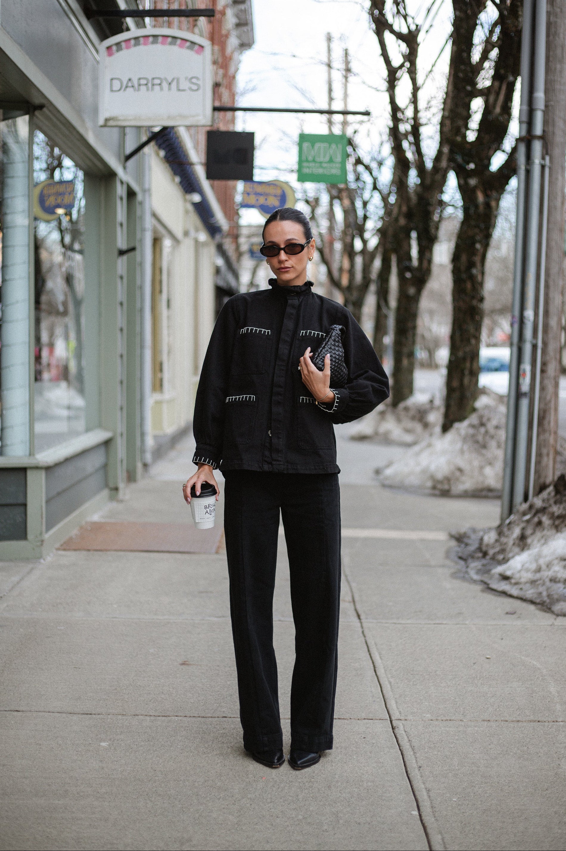 A woman holding coffee and wearing a black denim jacket with a frill collar and our signature white blanket stitching on four large patch pockets, paired with black jeans outdoors