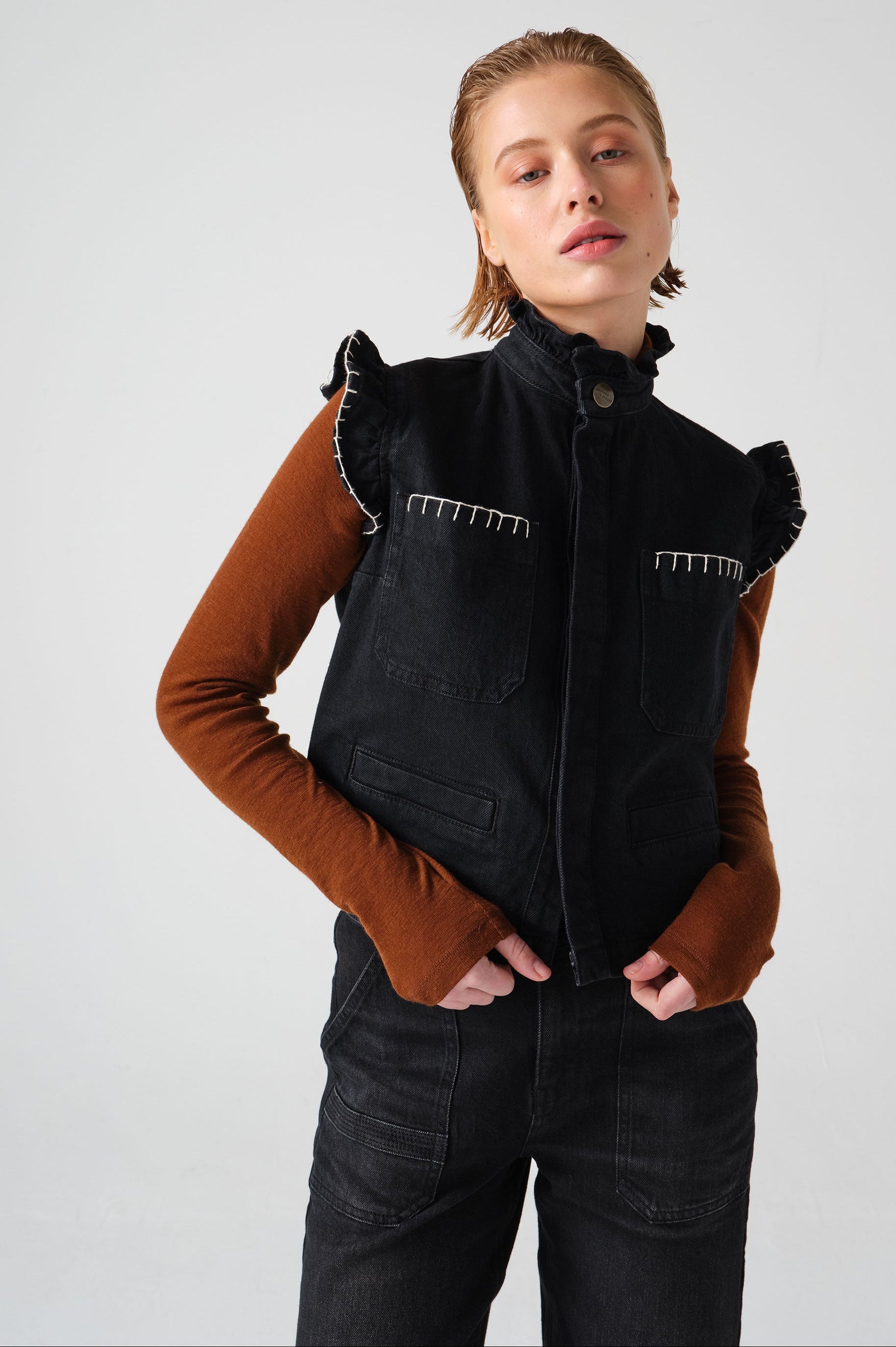 woman wearing a black vest/ waistcoat with a frill collar and our signature white stitching on two large patch pockets, paired with a brown top and black jeans against a white backdrop