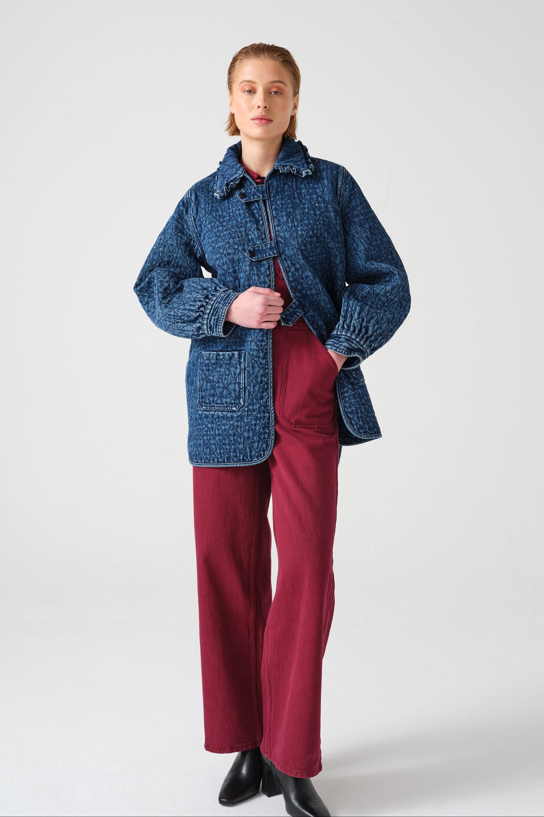 woman wearing a blue quilted jacket with a frill collar, tab closures, and two large pockets with red overalls on a white background