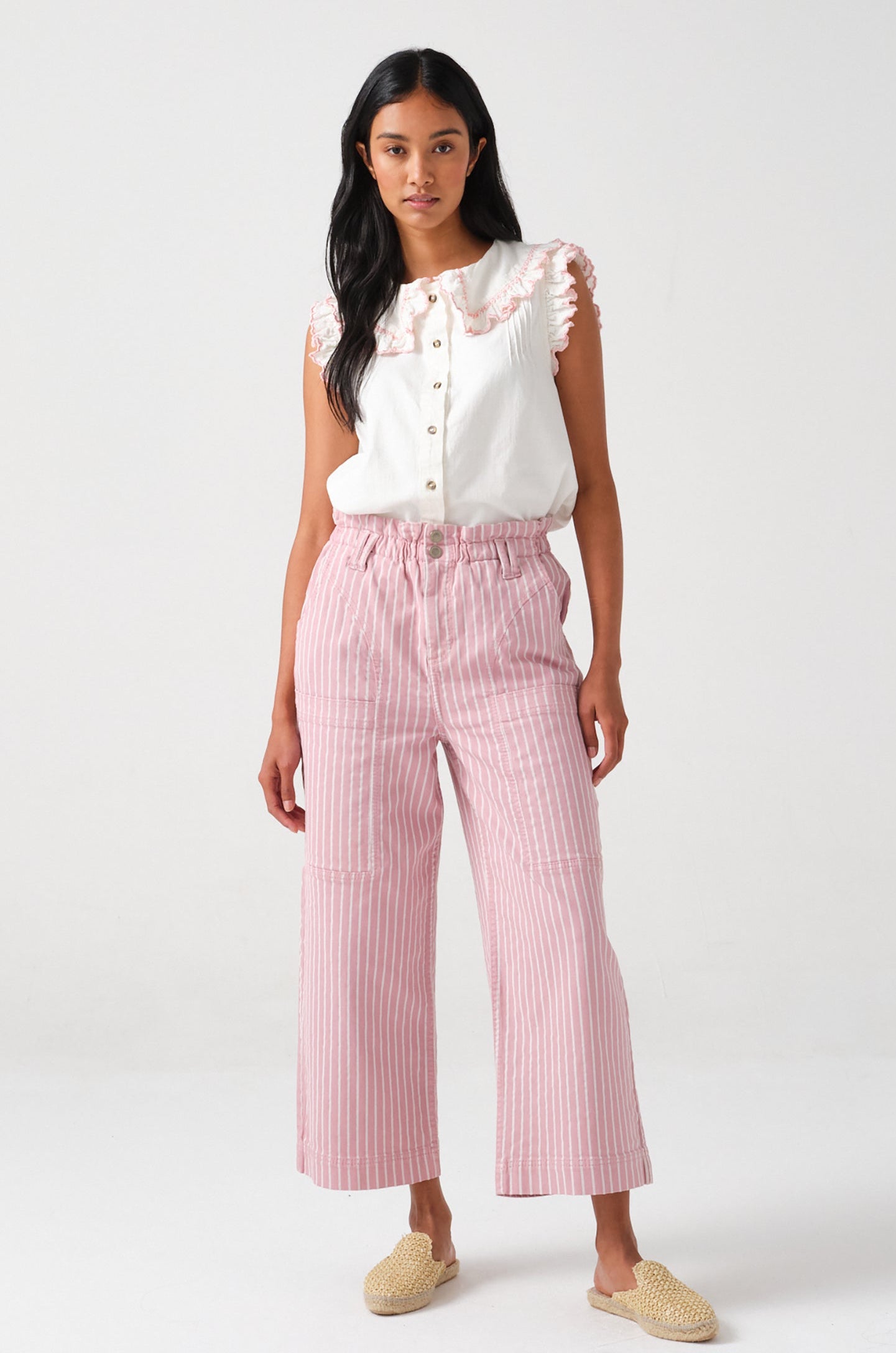 A woman wearing a short sleeve blouse with a frill collar and embroidered details in ecru and dusty rose colors, paired with striped pants.