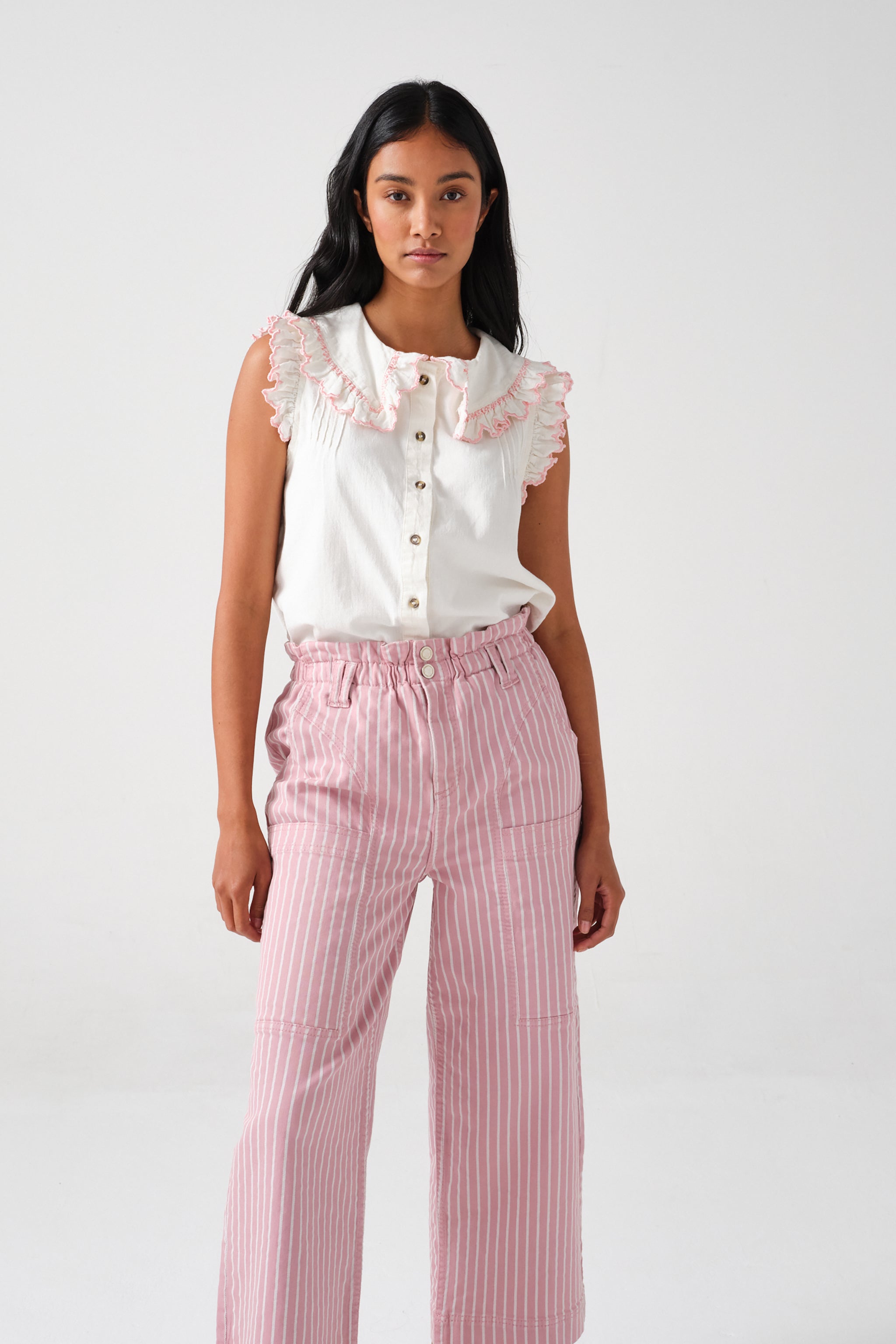 A woman wearing a short sleeve blouse with a frill collar and embroidered details in ecru and dusty rose colors, paired with striped pants.
