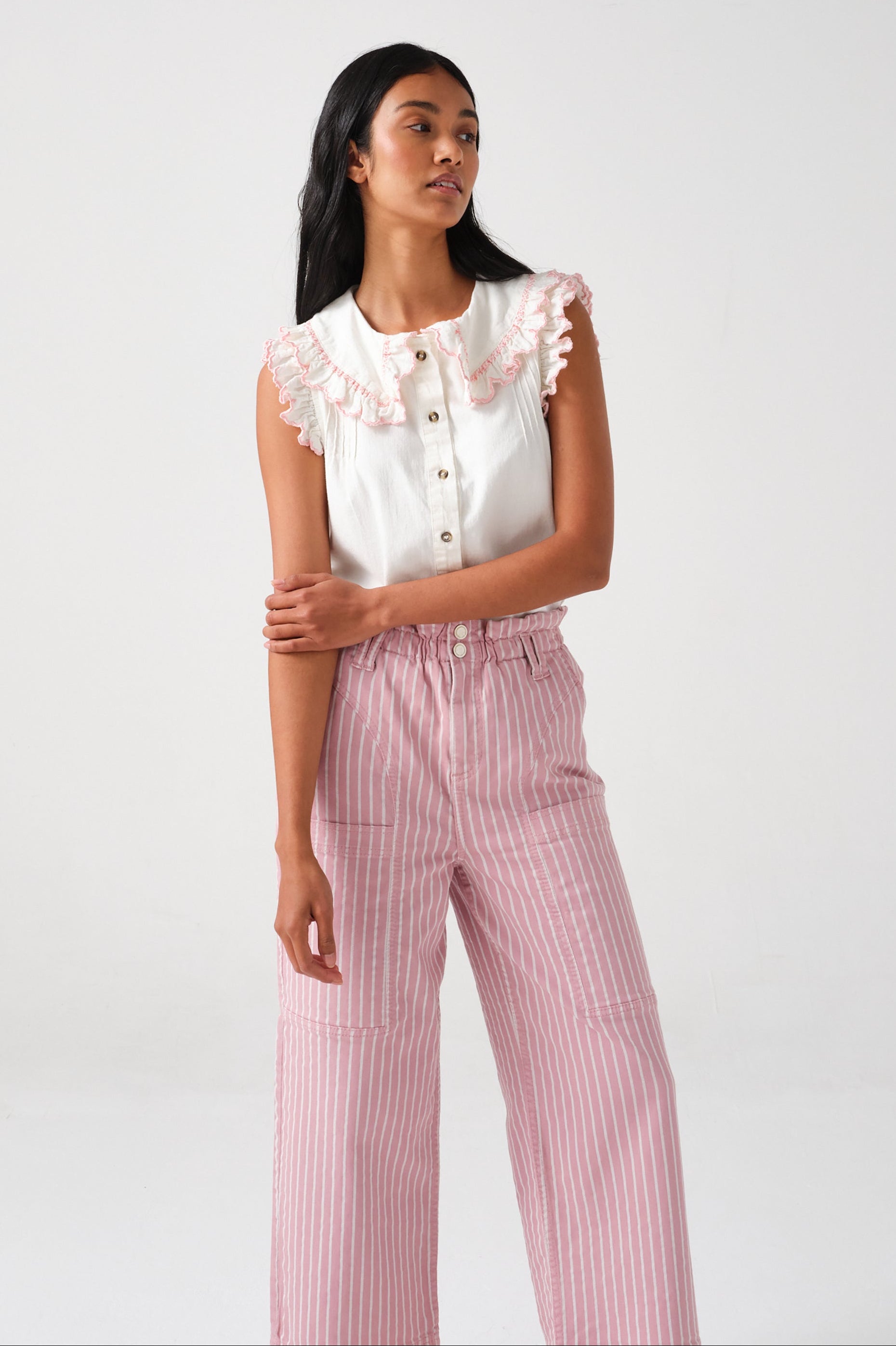 A woman wearing a short sleeve blouse with a frill collar and embroidered details in ecru and dusty rose colors, paired with striped pants.