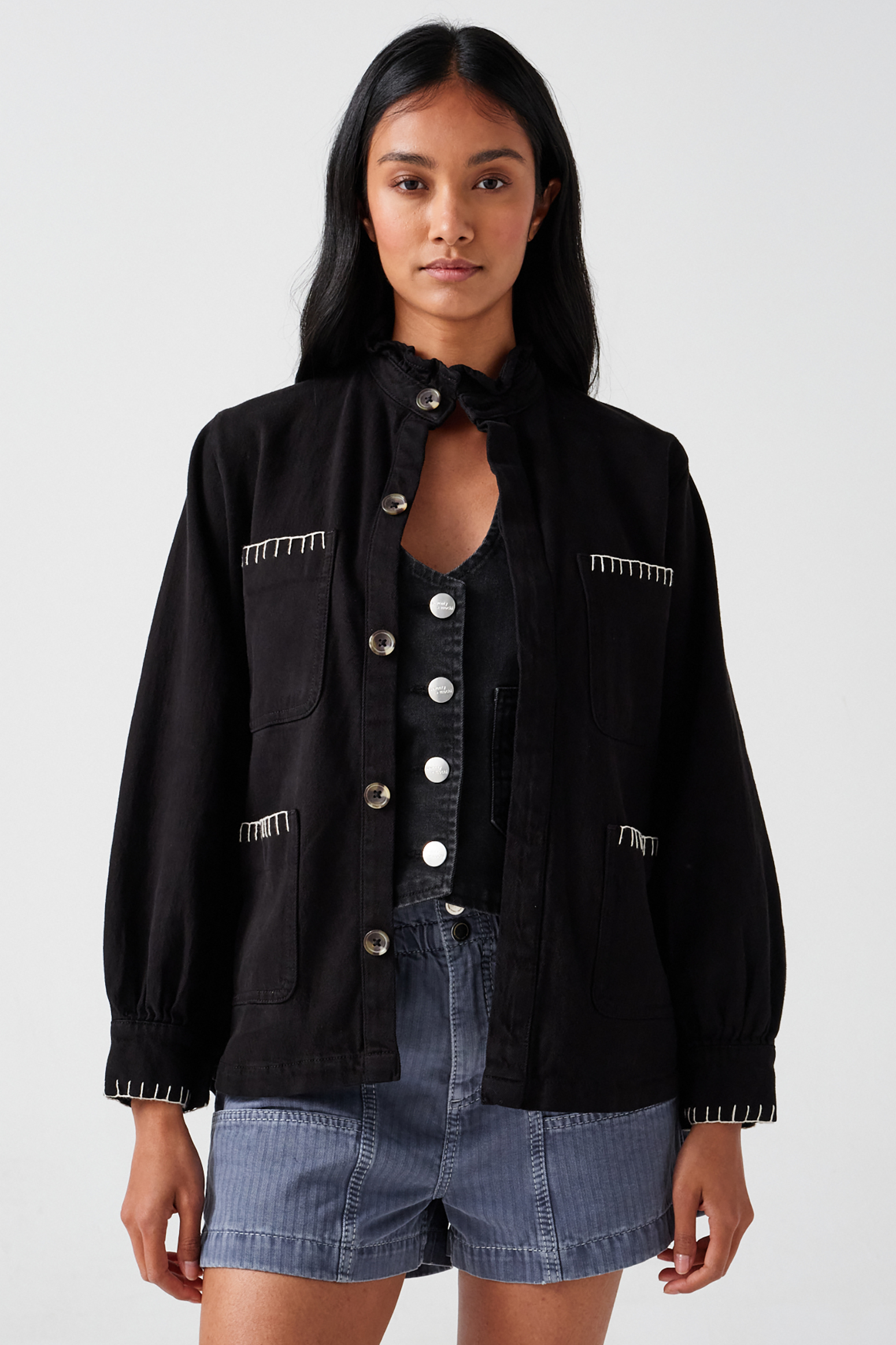 A woman wearing a black denim jacket with a frill collar and our signature white blanket stitching on four large patch pockets, paired with blue shorts and a black vest against a white backdrop
