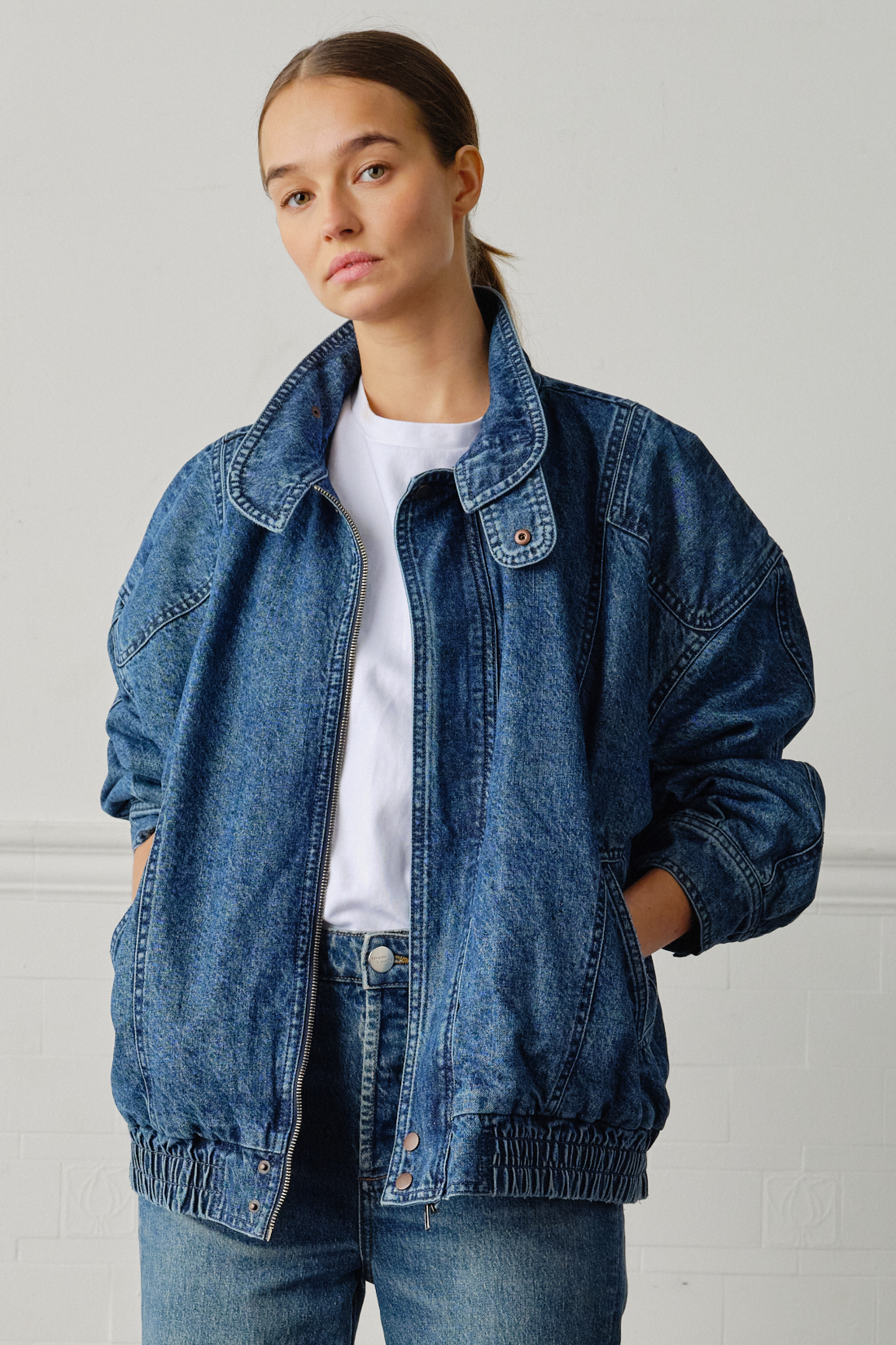 A woman wearing an oversized padded and seamed blue denim retro style bomber jacket with a round neckline, zip, and button closures at the top, bottom, and collar with a white top and blue jeans against a white backdrop