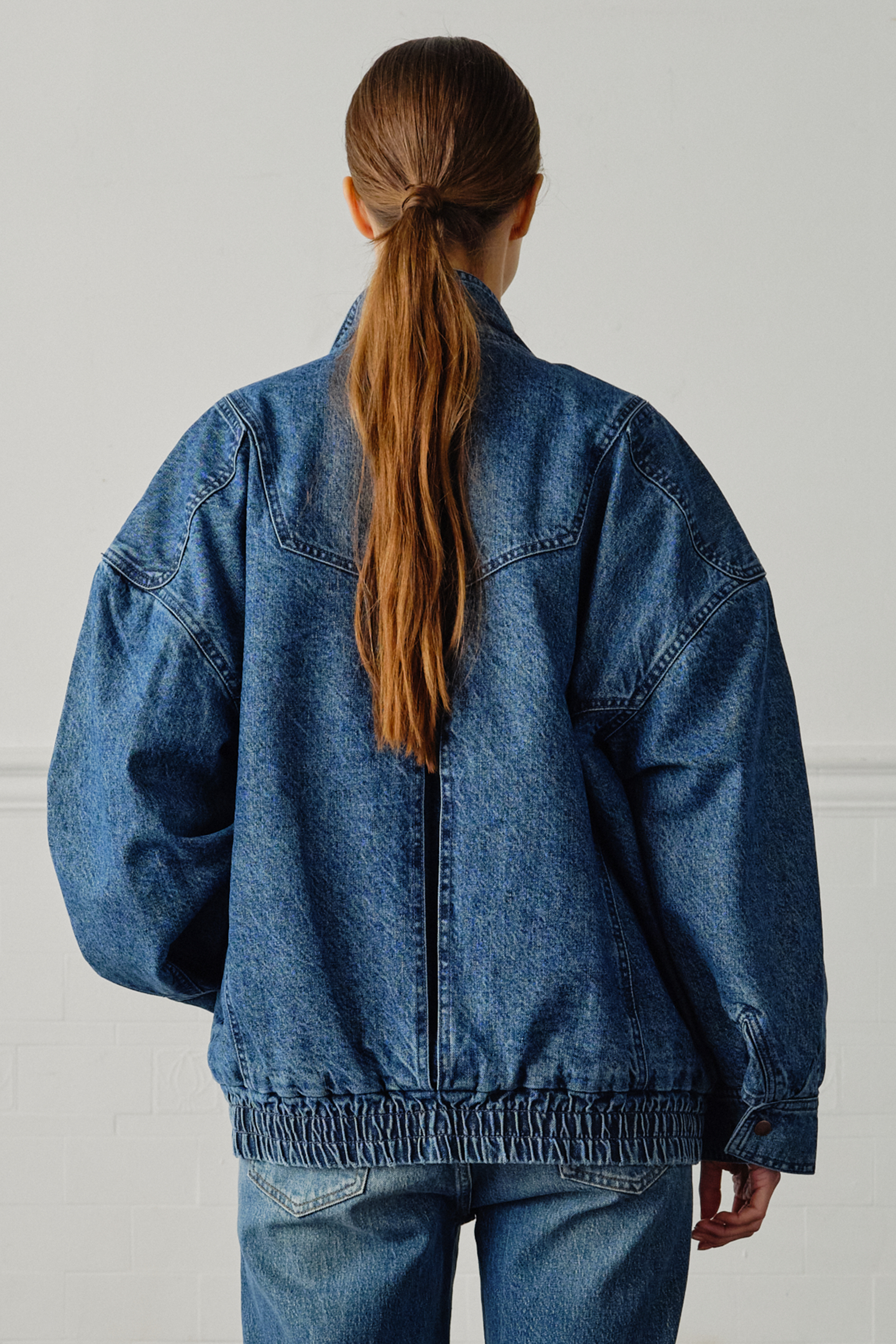 A woman wearing an oversized padded and seamed blue denim retro style bomber jacket with a round neckline, zip, and button closures at the top, bottom, and collar with a white top and blue jeans against a white backdrop
