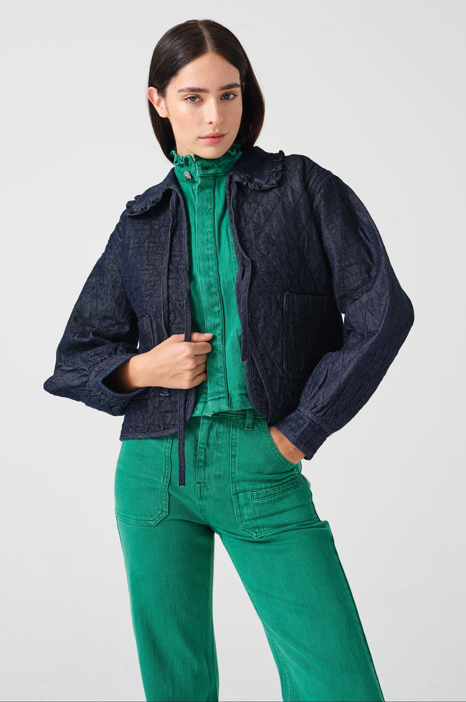 Woman wearing a dark blue denim quilted jacket with a slightly frilled collar and tie closures with a green denim shirt and green denim jeans
