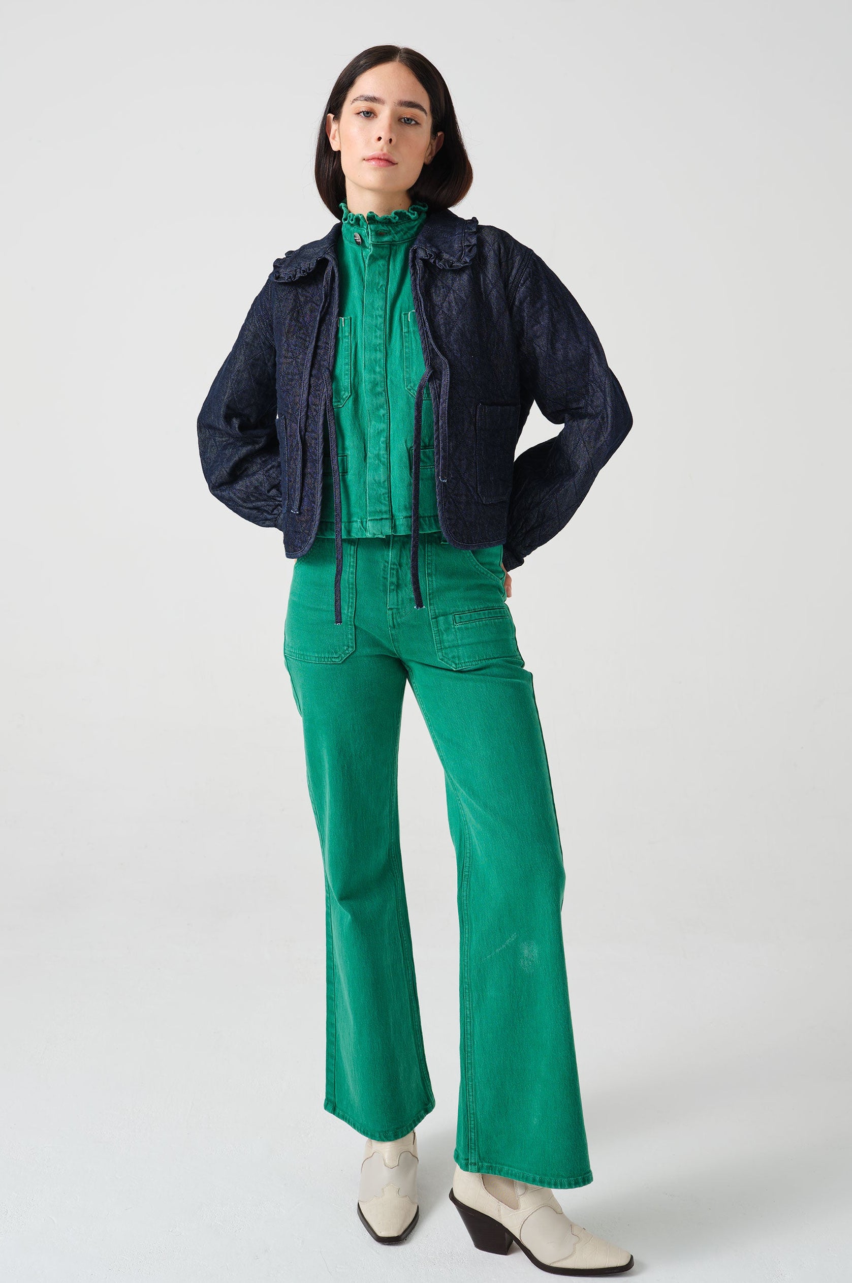 Woman wearing a dark blue denim quilted jacket with a slightly frilled collar and tie closures with a green denim shirt and green denim jeans