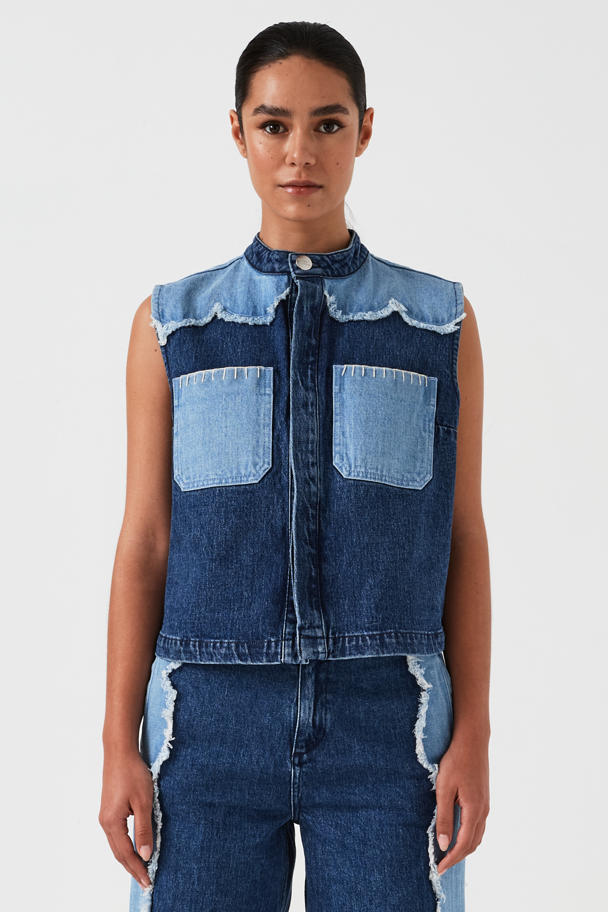 woman wearing a dark and light blue denim vest/ waistcoat with scallop frayed details and our signature white blanket stitching on two large patch pockets with blue jeans with scallop frayed details against a white backdrop