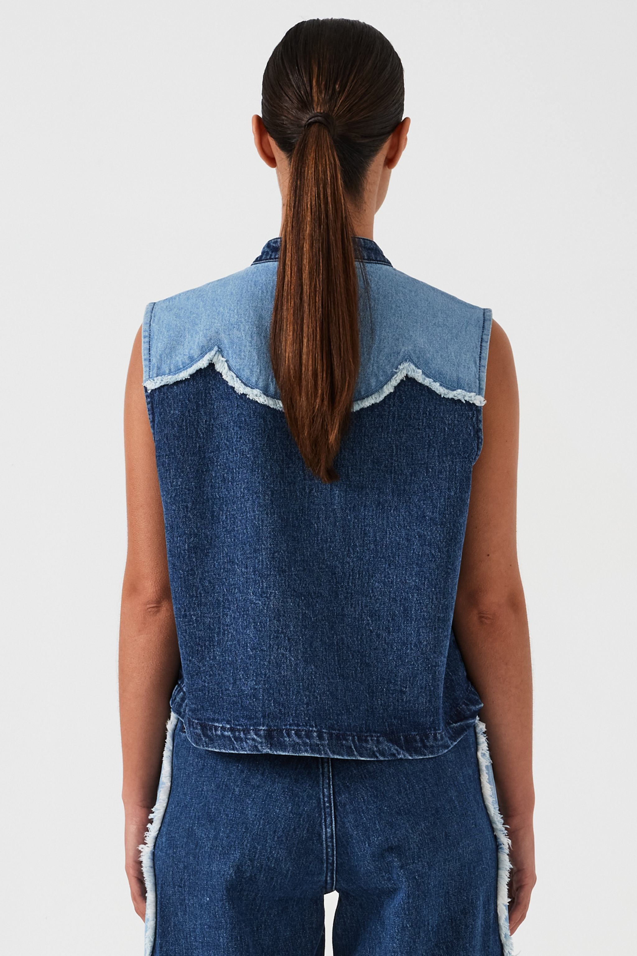woman wearing a dark and light blue denim vest/ waistcoat with scallop frayed details and our signature white blanket stitching on two large patch pockets with blue jeans with scallop frayed details against a white backdrop