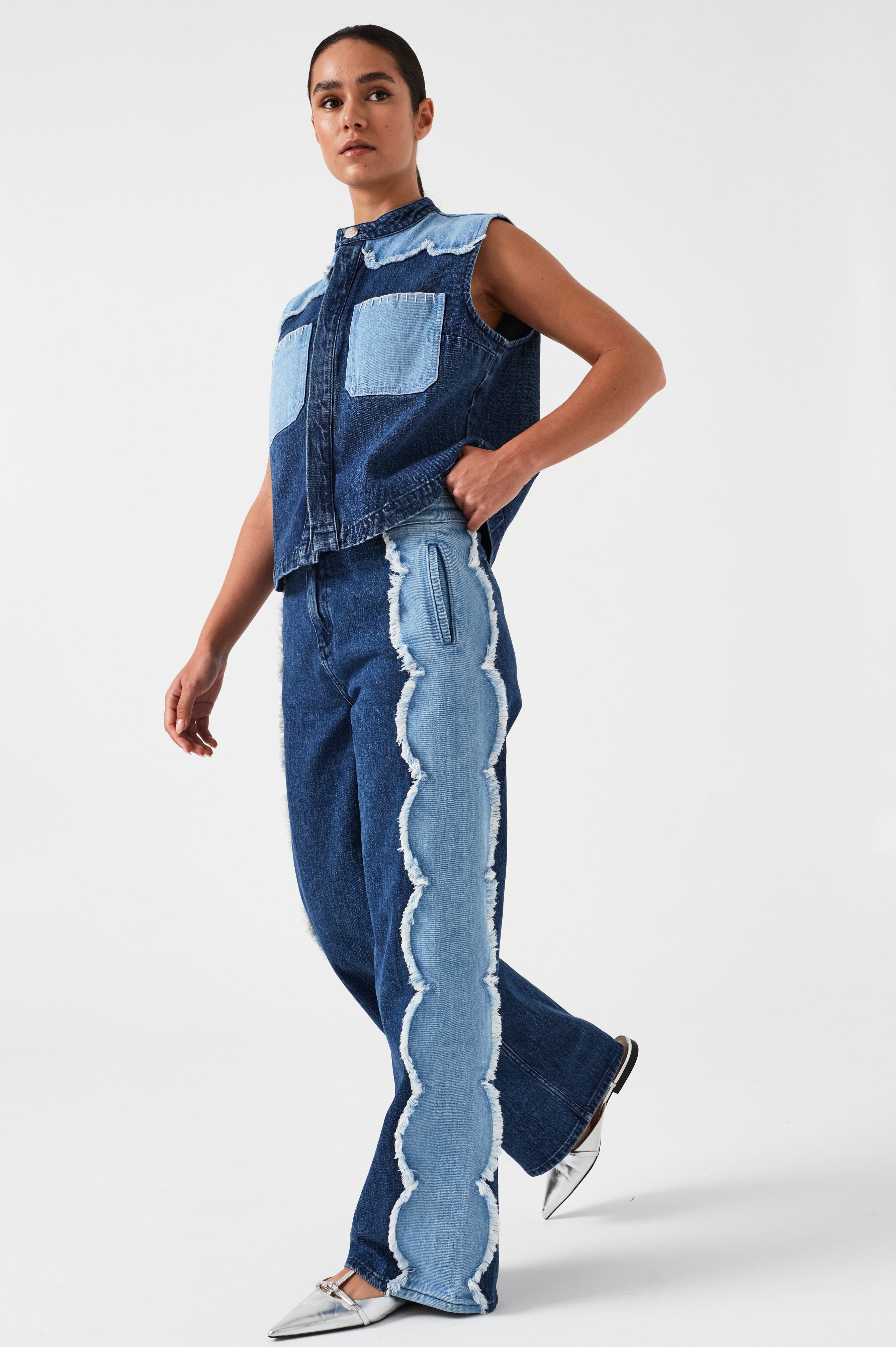 woman wearing a dark and light blue denim vest/ waistcoat with scallop frayed details and our signature white blanket stitching on two large patch pockets with blue jeans with scallop frayed details against a white backdrop