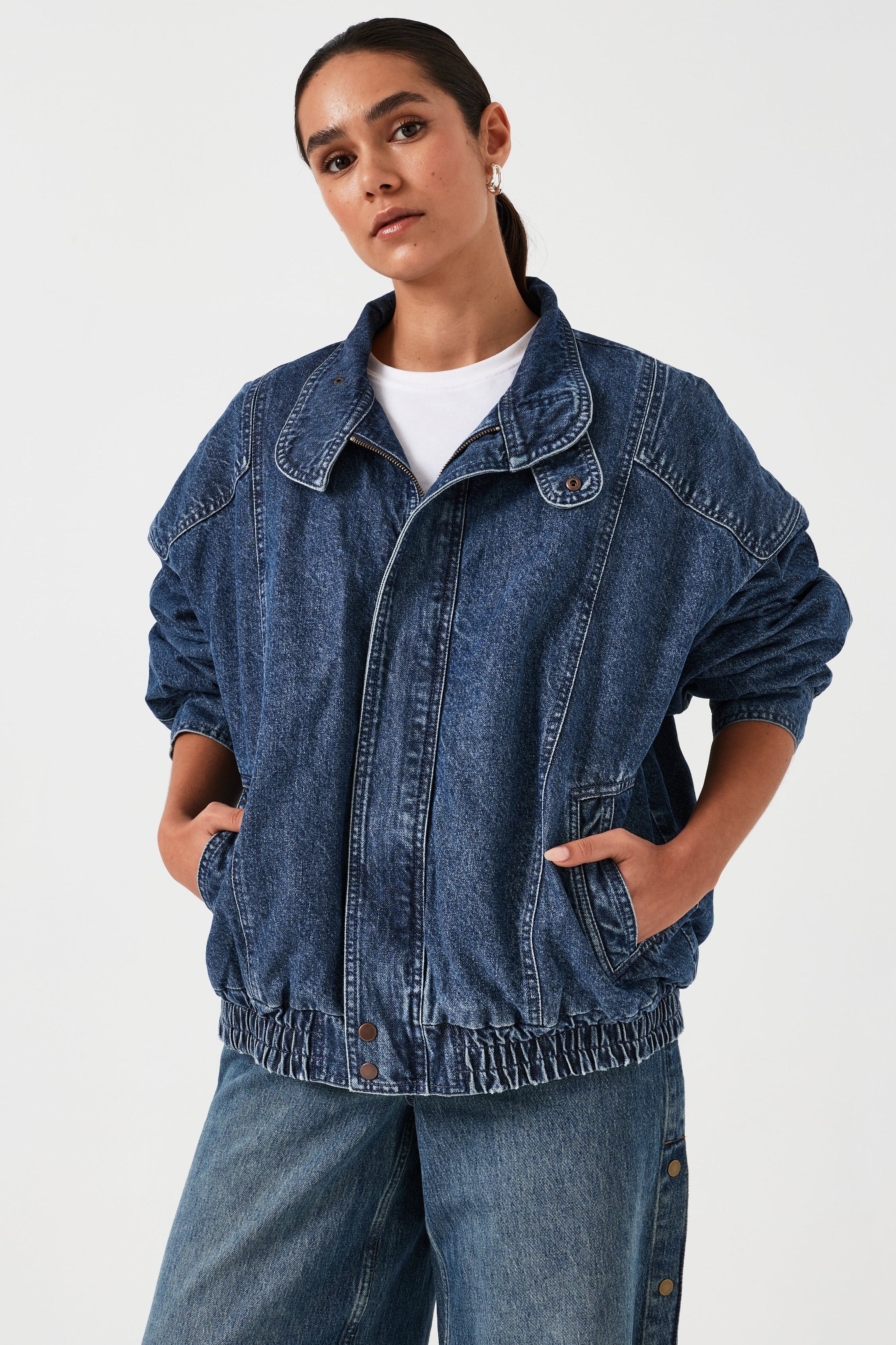 A woman wearing an oversized padded and seamed blue denim retro style bomber jacket with a round neckline, zip, and button closures at the top, bottom, and collar with a white top and blue jeans against a white backdrop