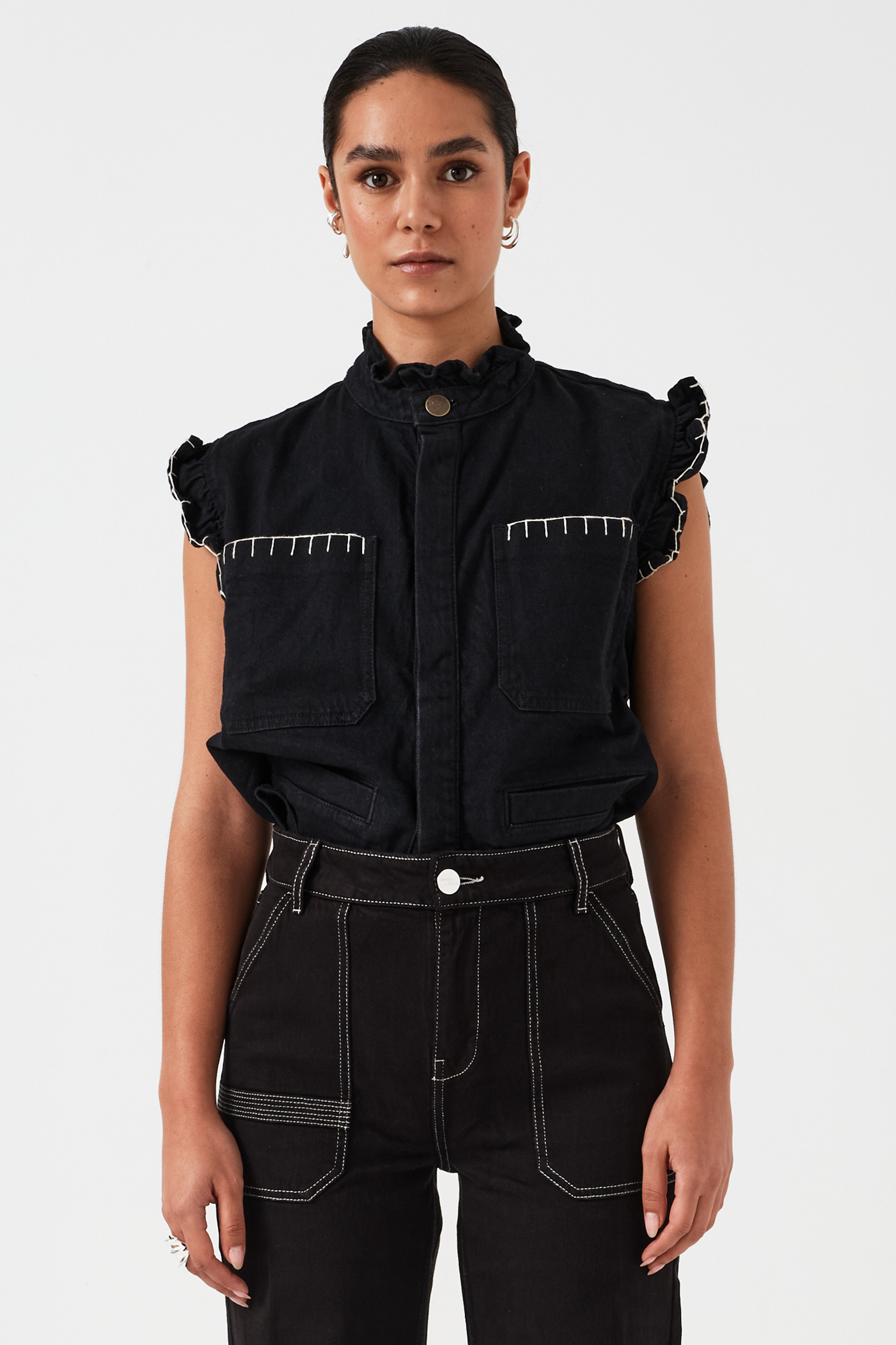 woman wearing a black vest/ waistcoat with a frill collar and our signature white stitching on two large patch pockets, paired with black jeans against a white backdrop