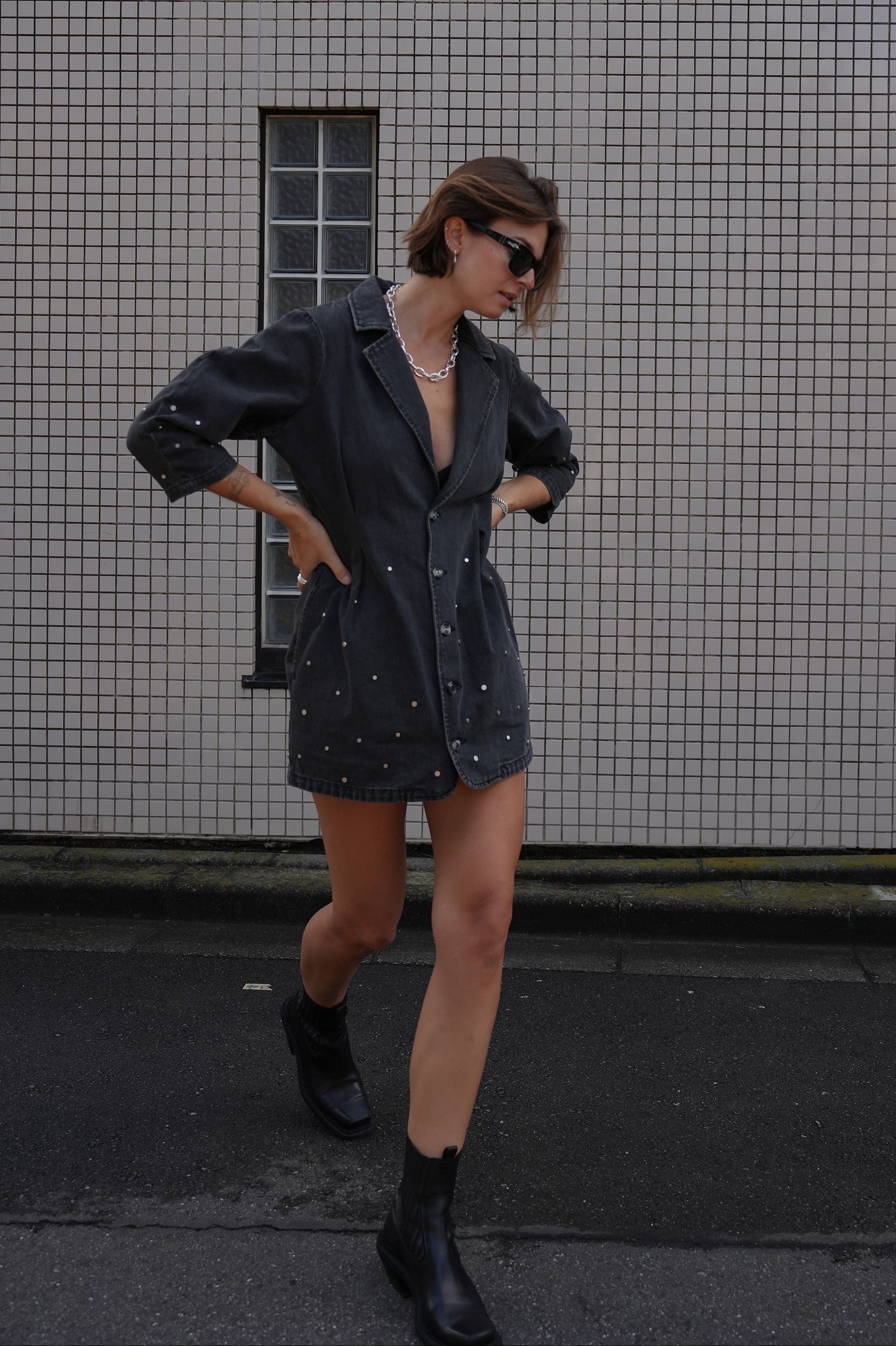 Woman wearing a black denim dress with silver studs (embellishments) and a tailored cinched waist against a tiled wall.