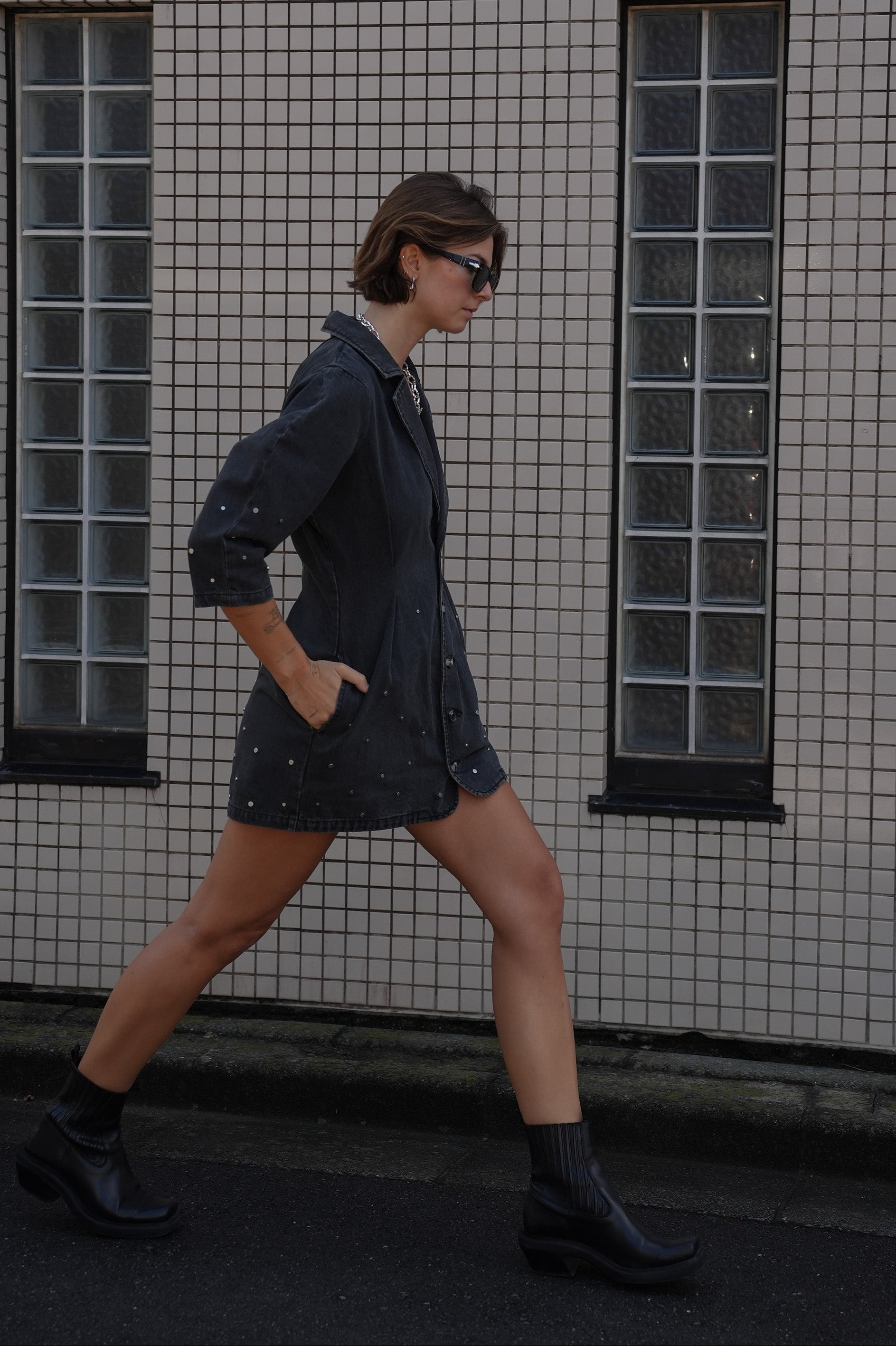 Woman wearing a black denim dress with silver studs (embellishments) and a tailored cinched waist against a tiled wall.