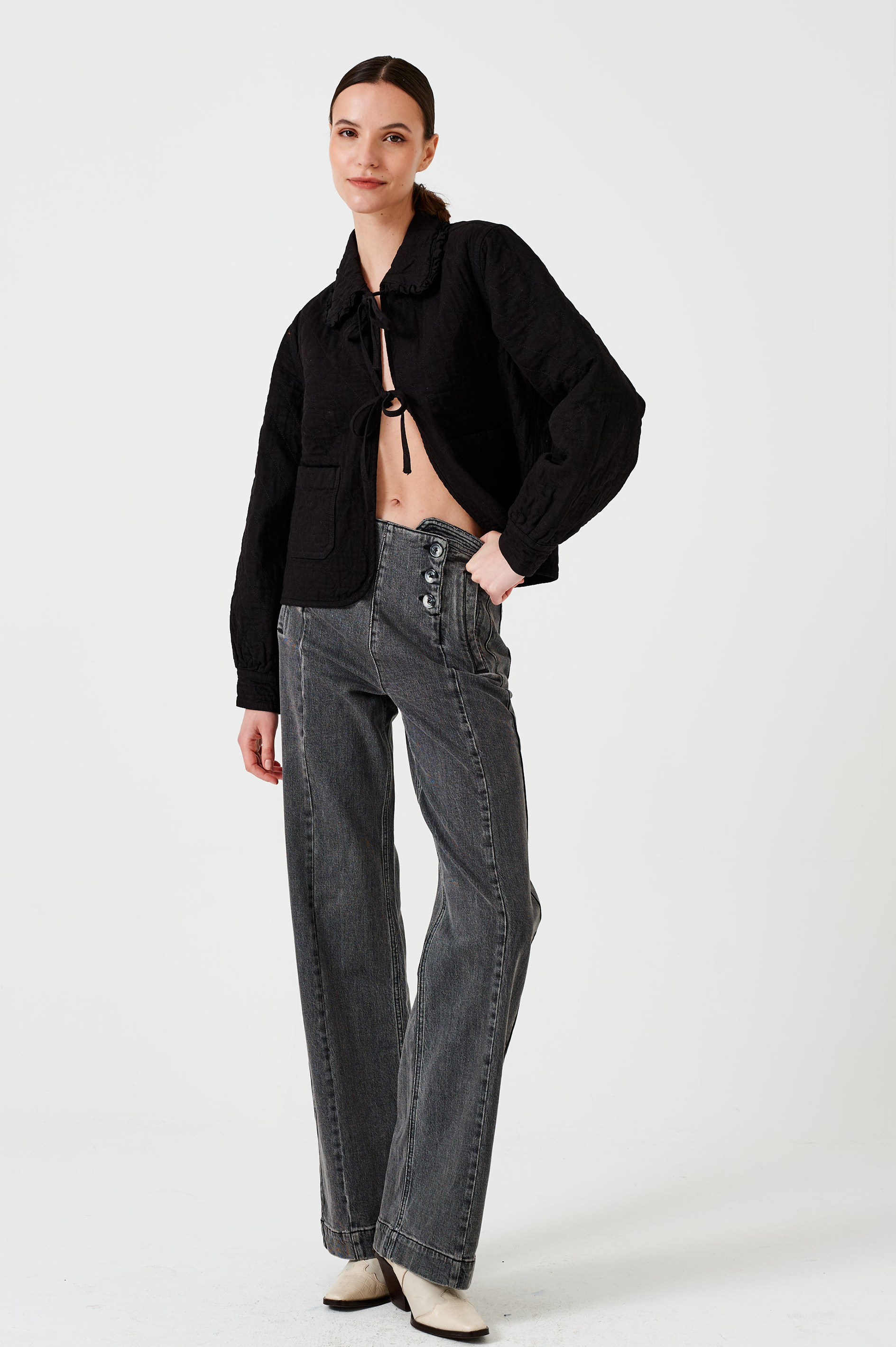Woman wearing a black quilted denim jacket with a ruffled collar, front tie closures, and two large pockets, and black jeans on a white background