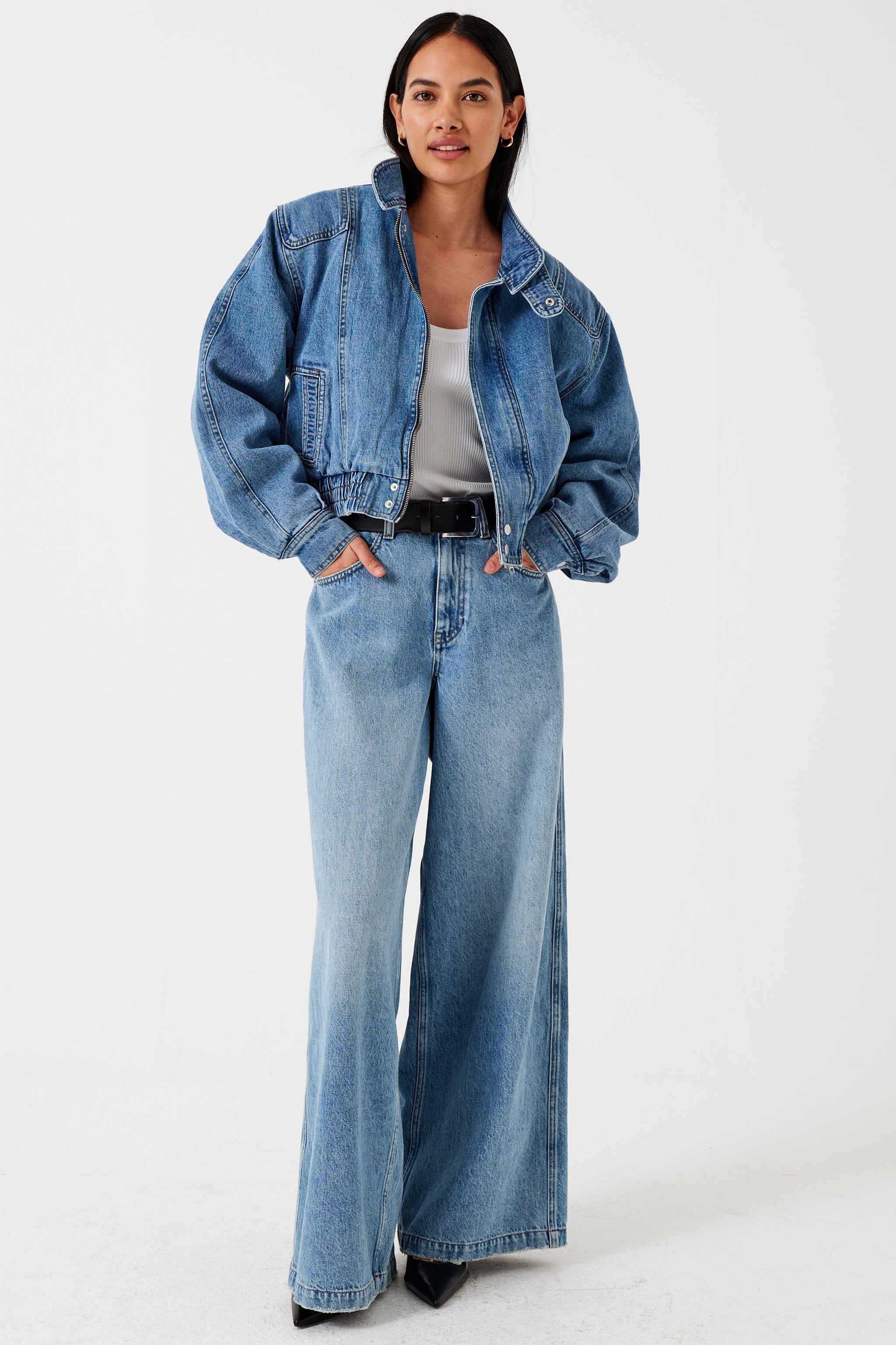 A woman wearing a high-rise, wide-leg, light wash blue denim jean, white shirt, and denim bomber jacket with a belt and black shoes against a white background