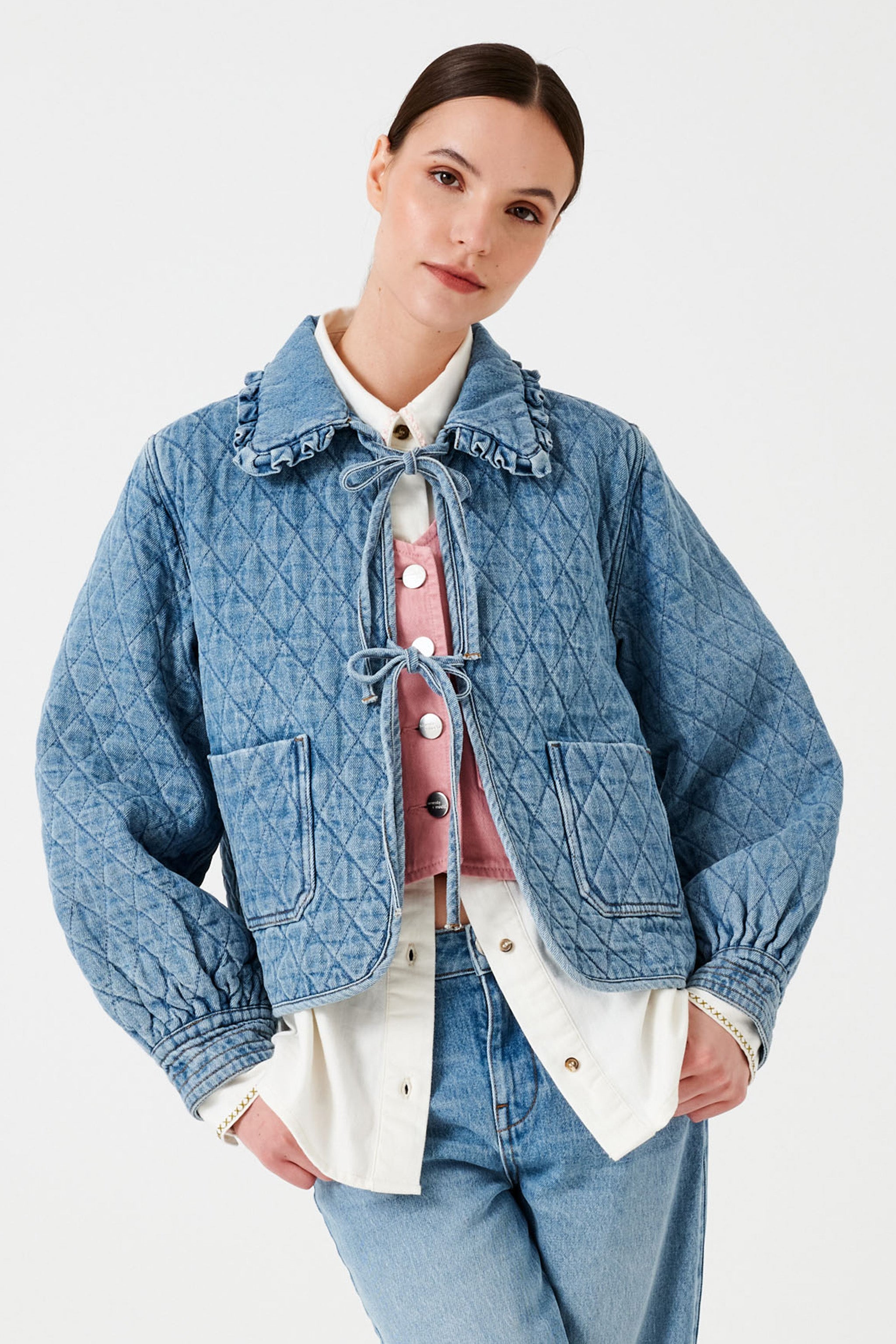 A woman wearing a light blue quilted denim jacket with a frill collar, front tie closures, and two large pockets paired with a white shirt, pink vest and blue jeans.