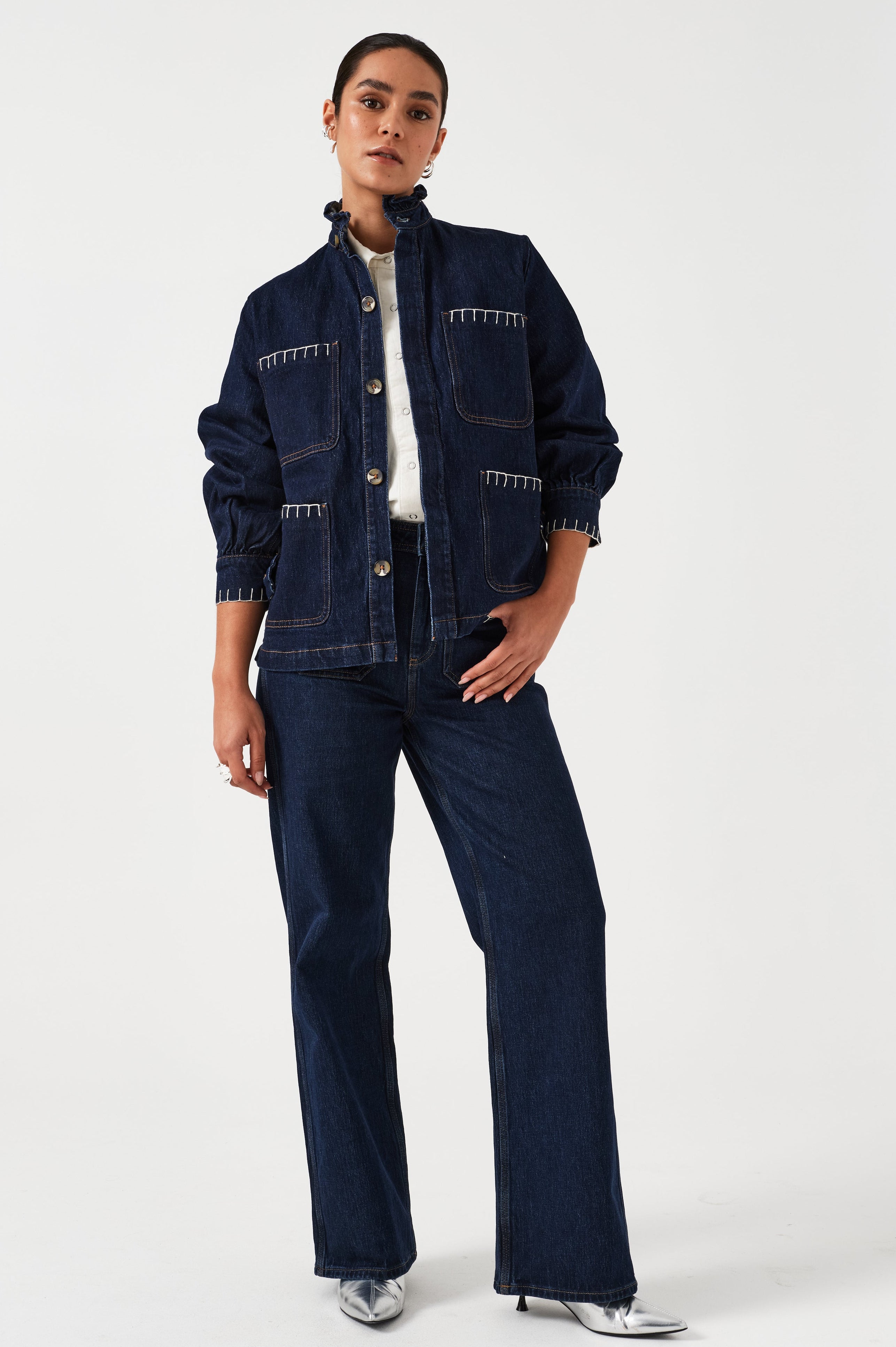 A woman wearing a dark blue vintage style jacket with a frill collar and our signature white blanket stitching on four large patch pockets, paired with a dark blue jeans and a white top against a white background
