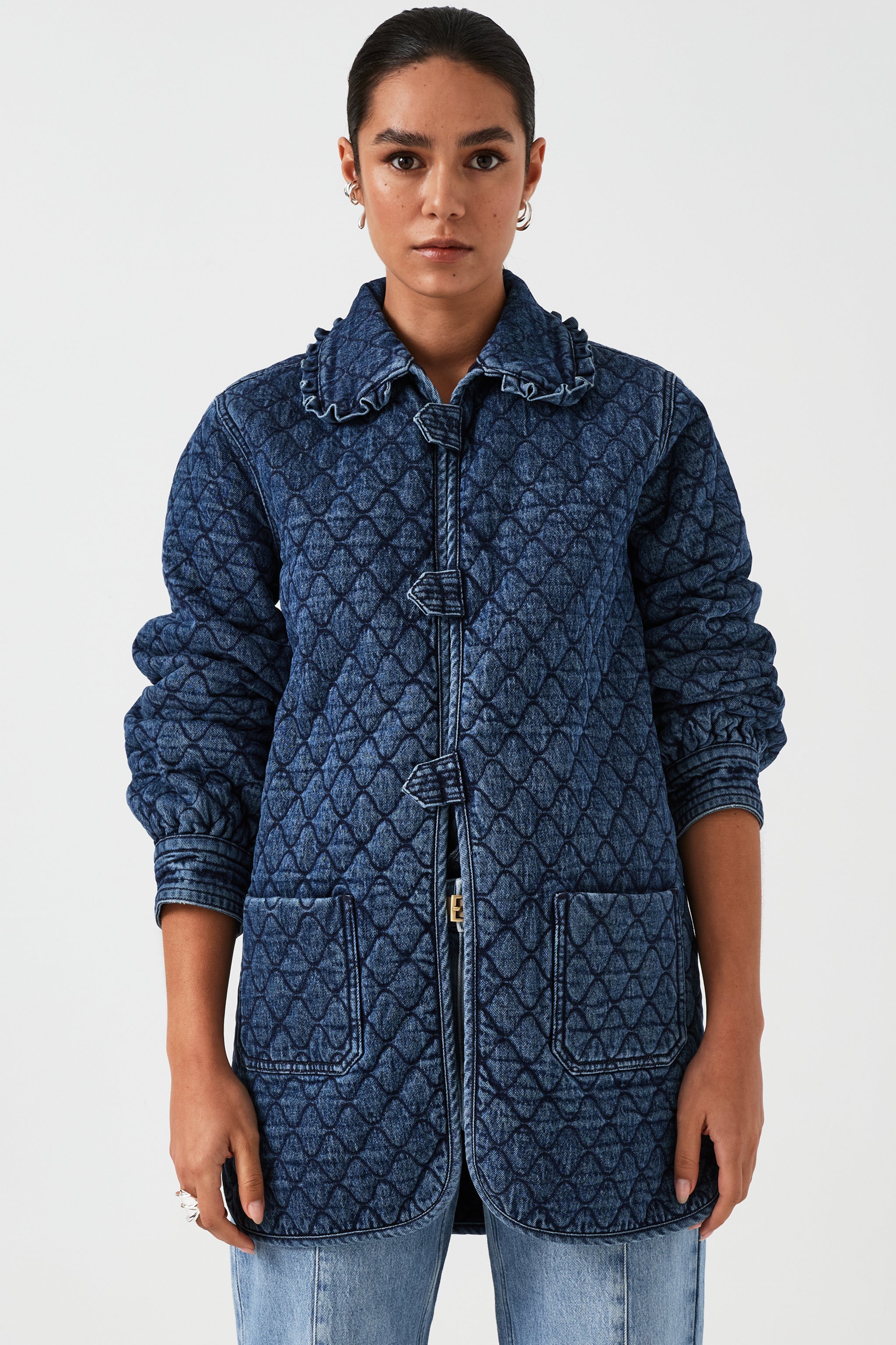 woman wearing a blue quilted jacket with a frill collar, tab closures, and two large pockets on a white background