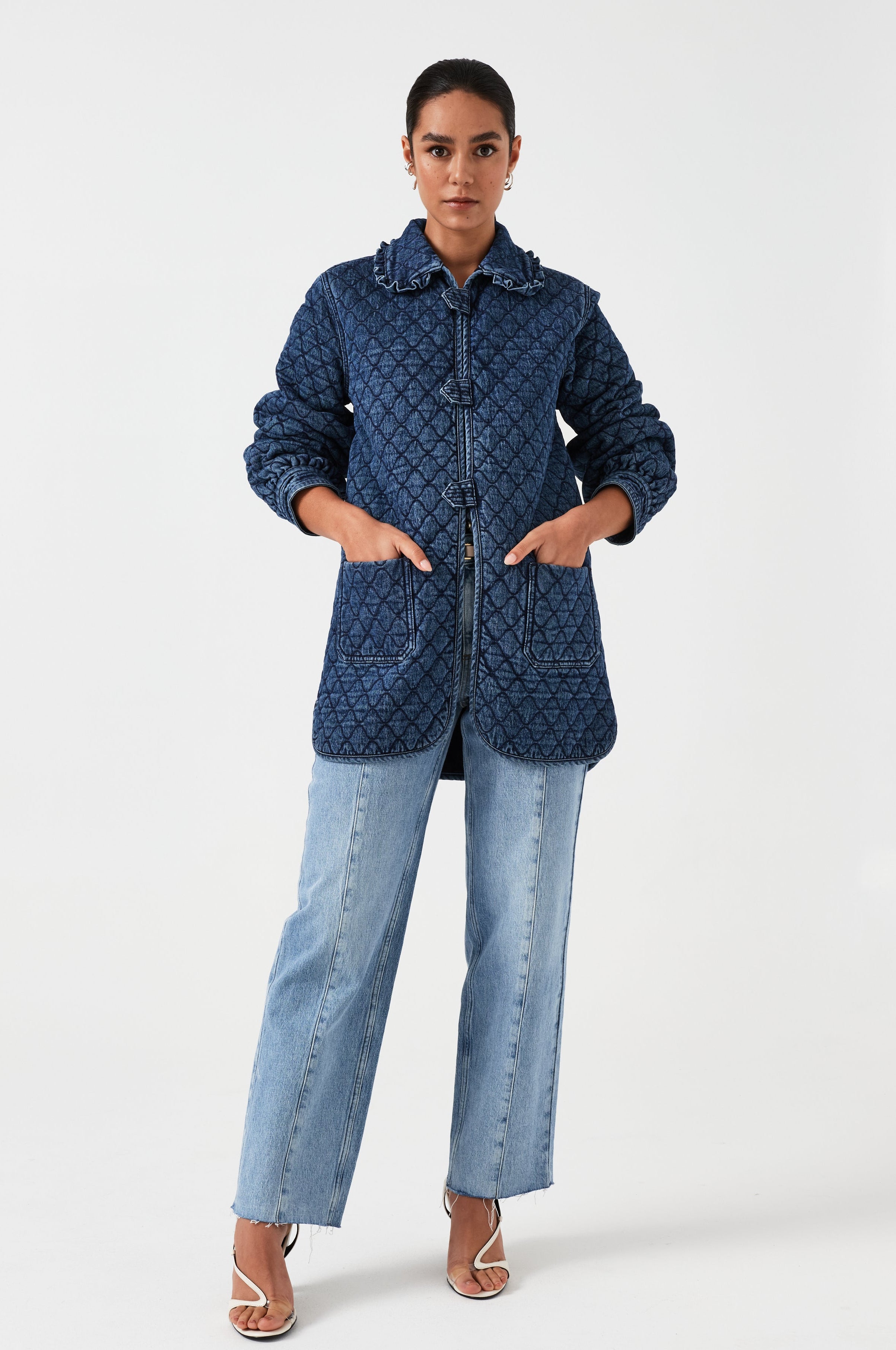 woman wearing a blue quilted jacket with a frill collar, tab closures, and two large pockets with blue jeans on a white background