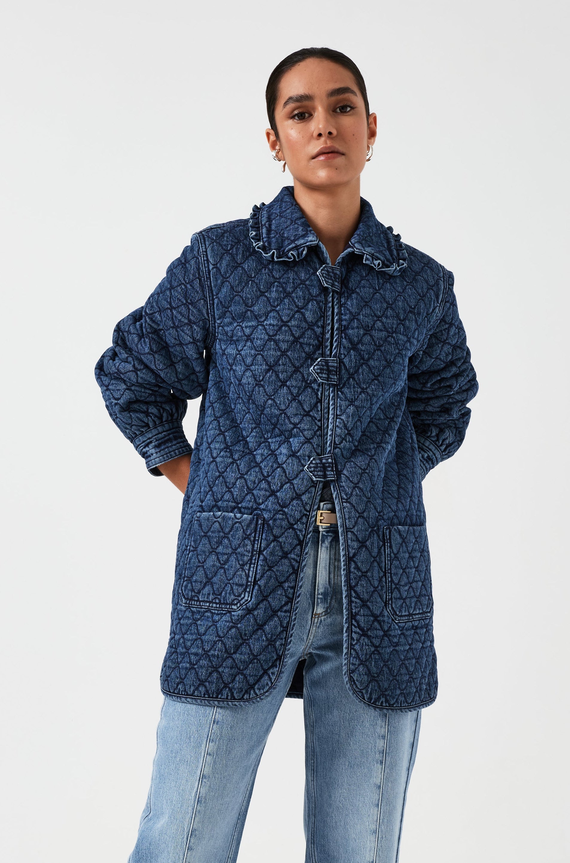 woman wearing a blue quilted jacket with a frill collar, tab closures, and two large pockets with blue jeans on a white background