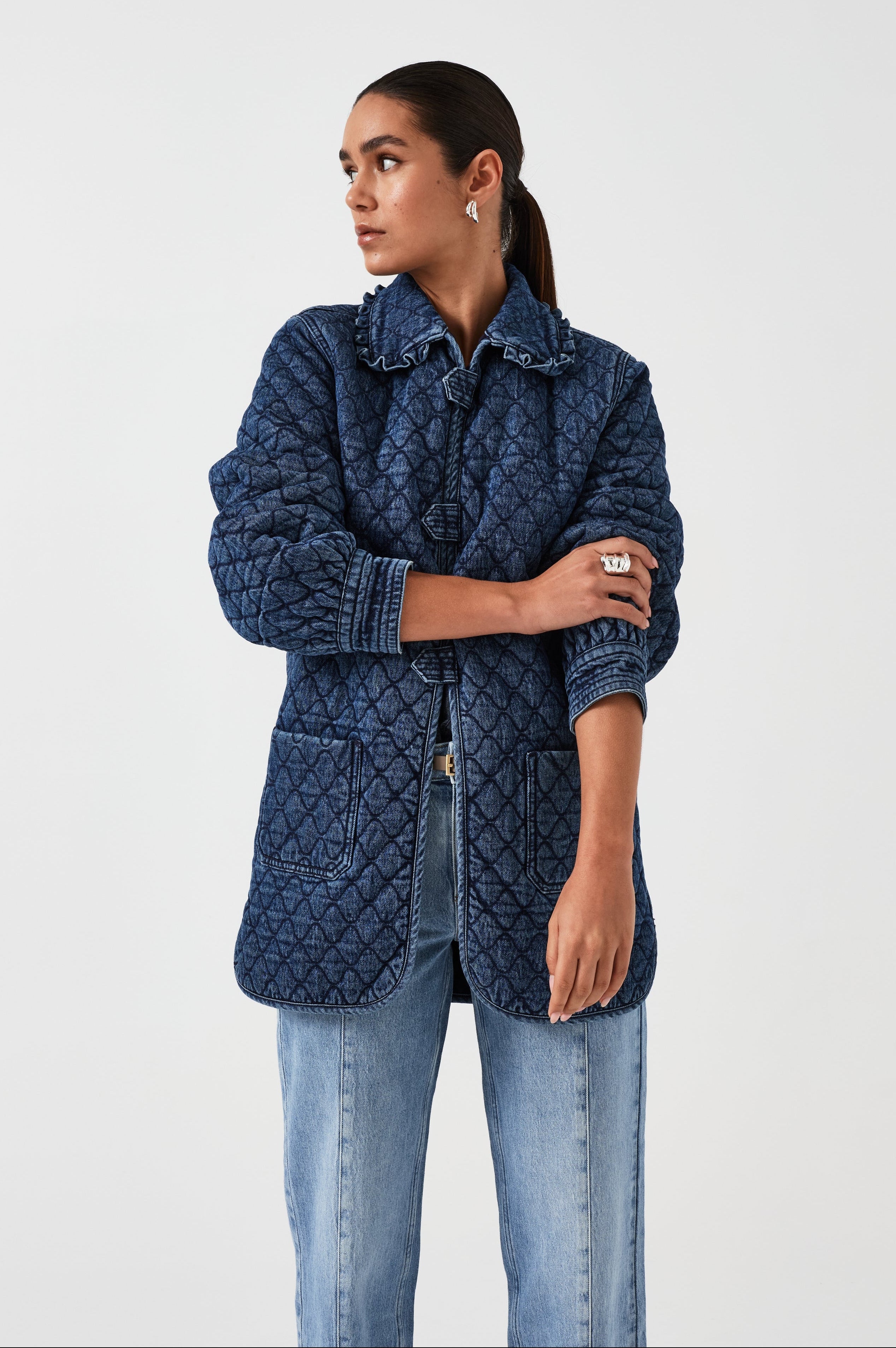 woman wearing a blue quilted jacket with a frill collar, tab closures, and two large pockets with blue jeans on a white background