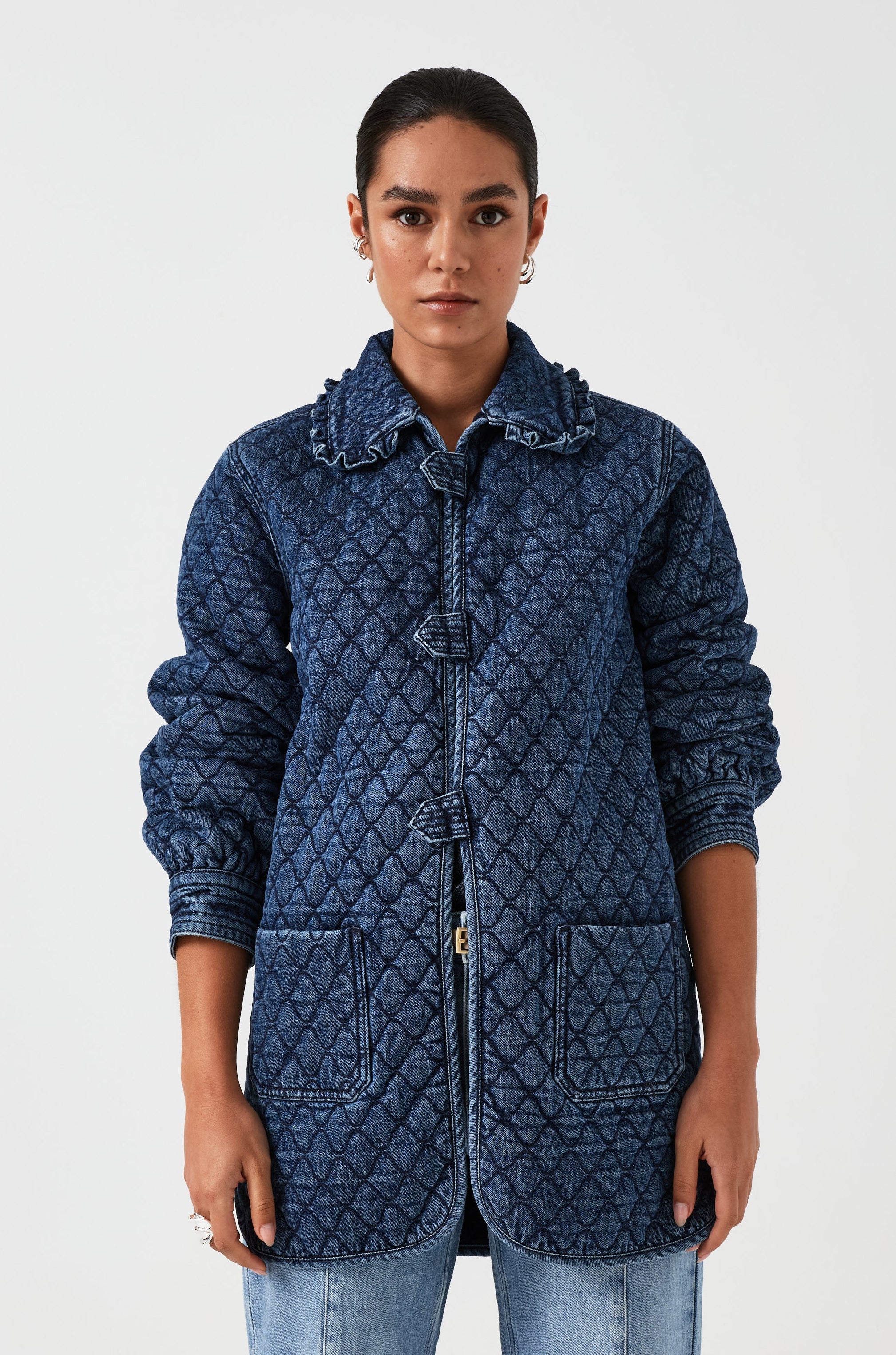 woman wearing a blue quilted jacket with a frill collar, tab closures, and two large pockets with blue jeans on a white background