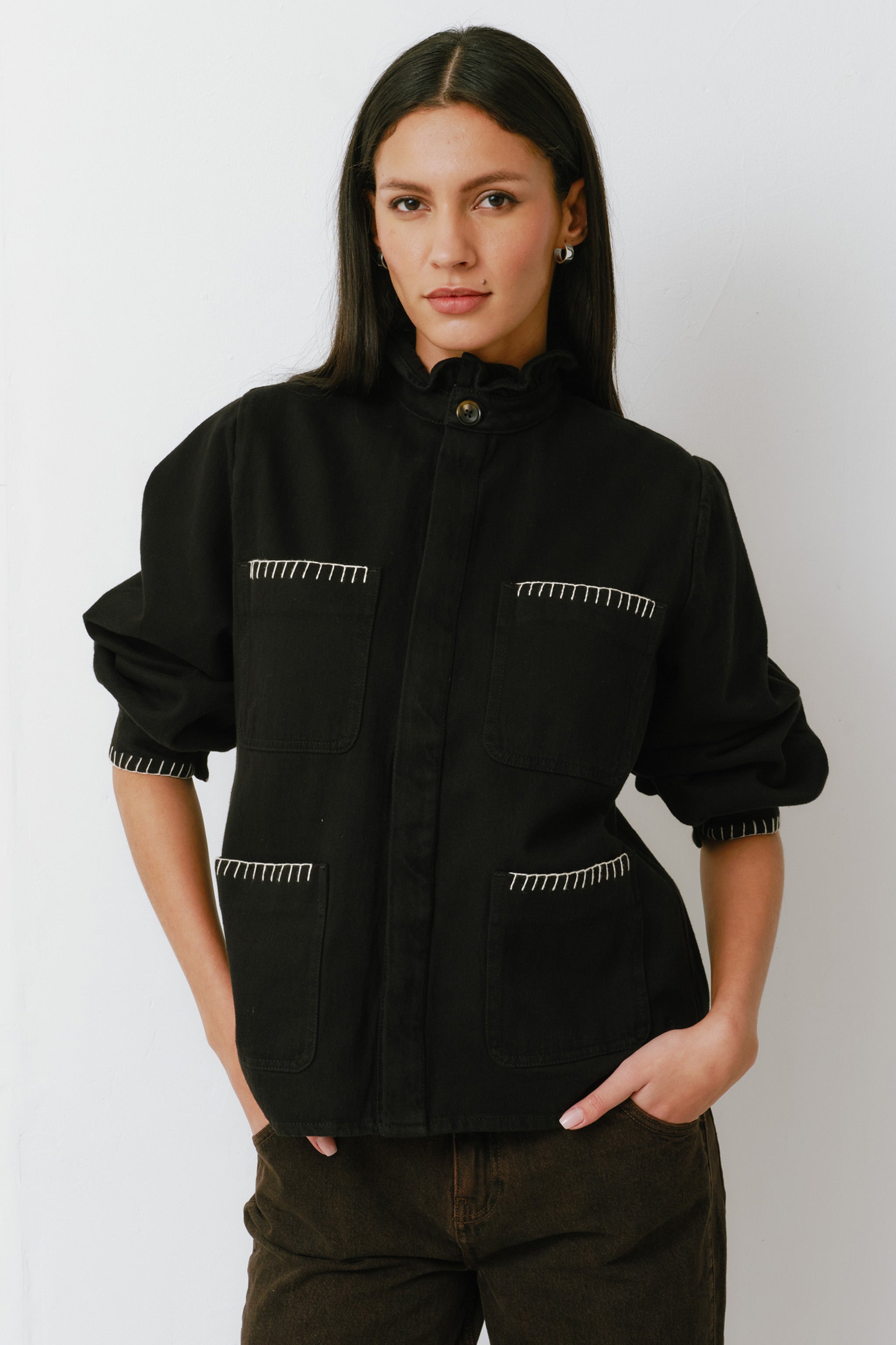 A woman wearing a black denim jacket with a frill collar and our signature white blanket stitching on four large patch pockets, paired with brown jeans against a white backdrop