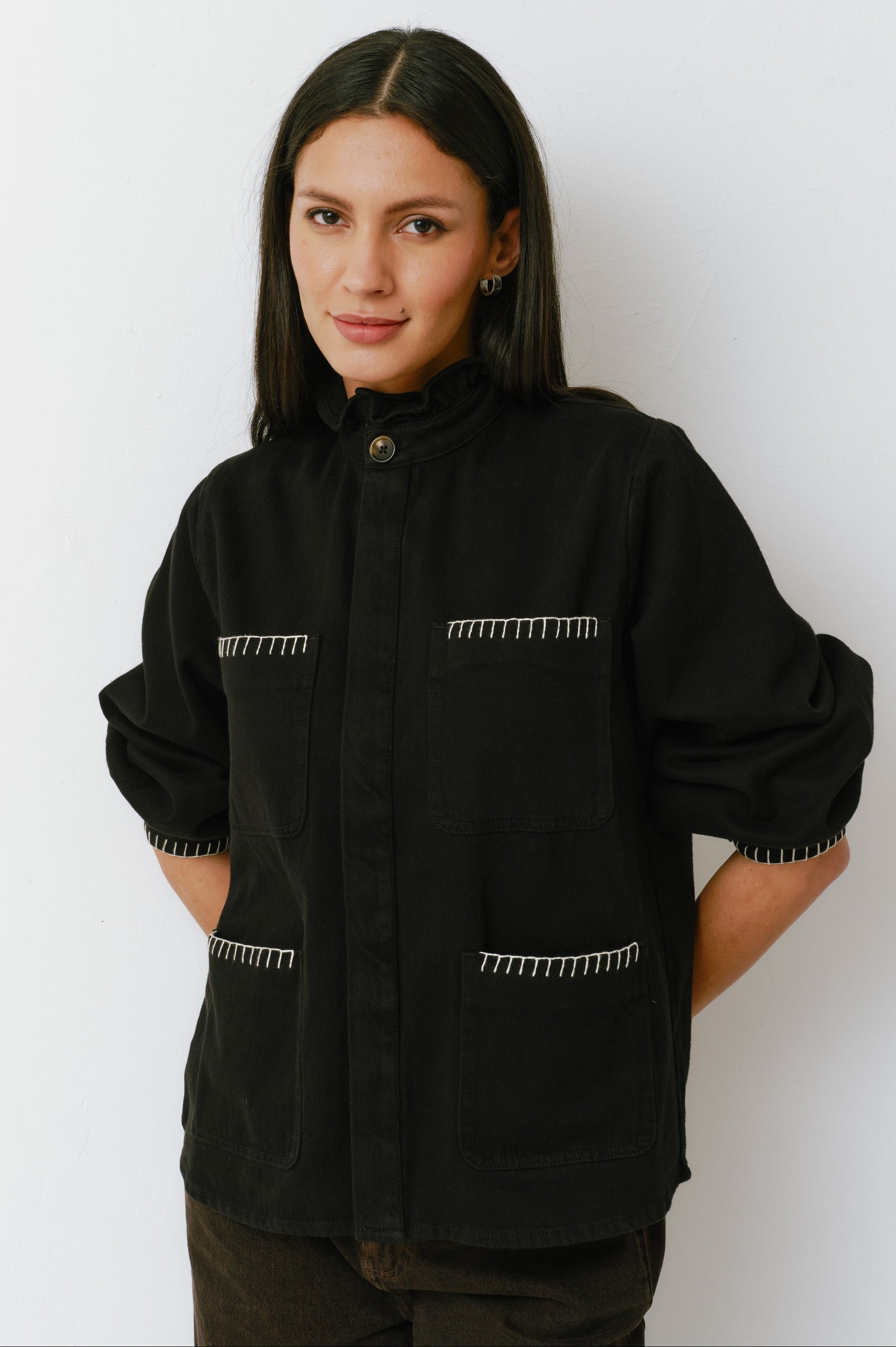 A woman wearing a black denim jacket with a frill collar and our signature white blanket stitching on four large patch pockets, paired with brown jeans against a white backdrop