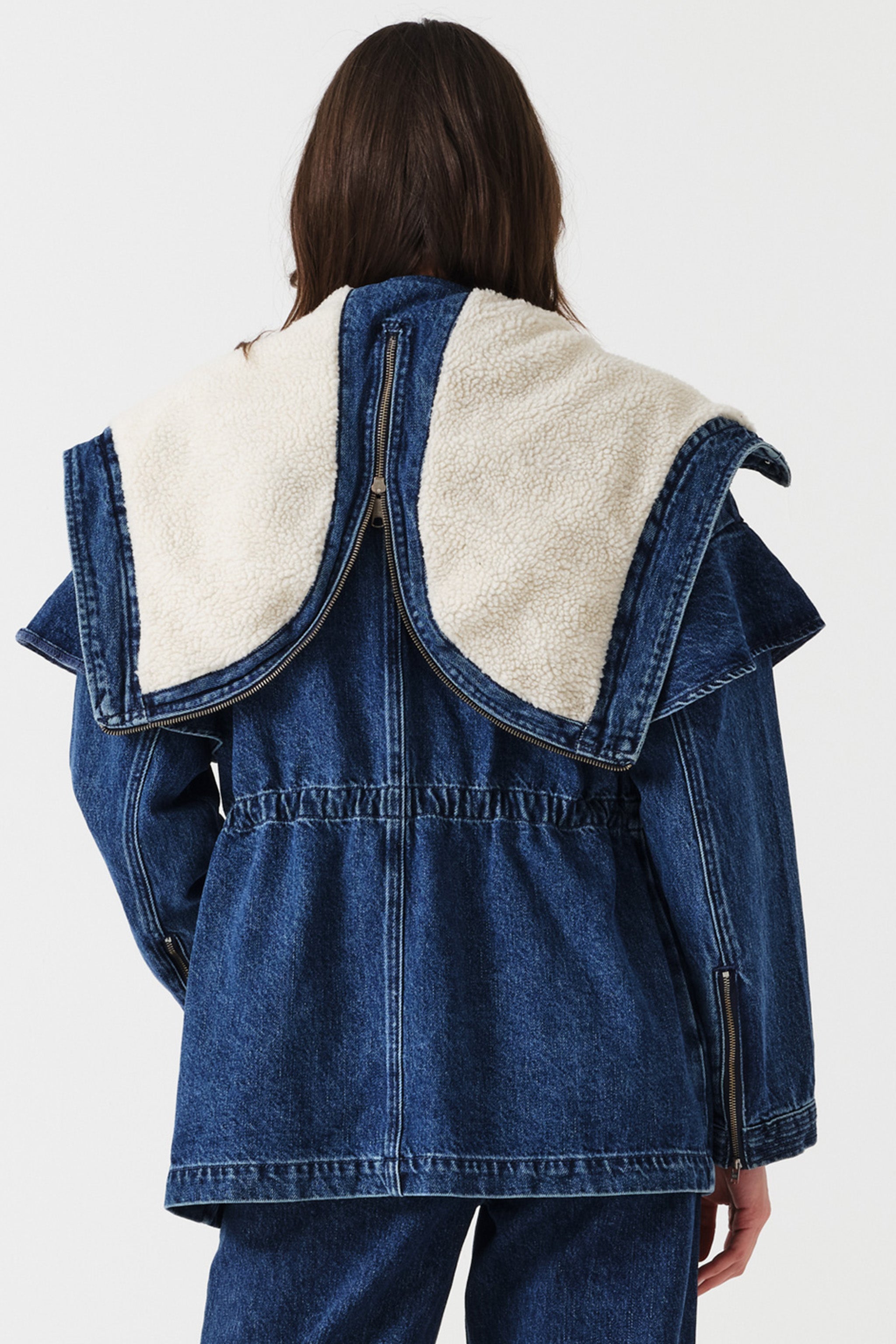 Woman wearing a dark blue denim jacket with a white sherpa lined removable hood, waist cinch, and zip pocket details on a white background, photographed from the back