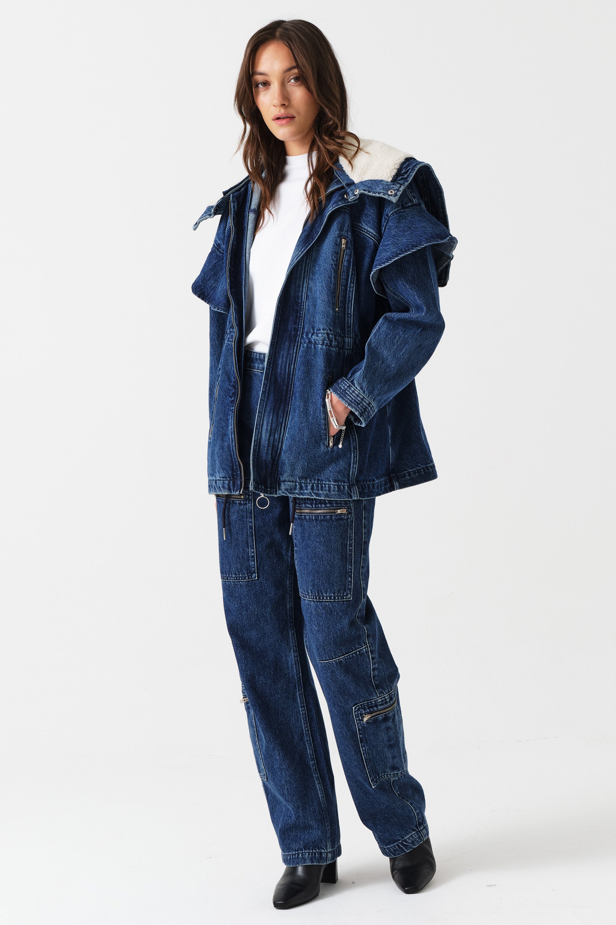 Woman wearing a white shirt, dark blue denim jacket with a white sherpa lined removable hood, waist cinch, and zip pocket details with dark blue jeans with cargo zip pockets on a white background