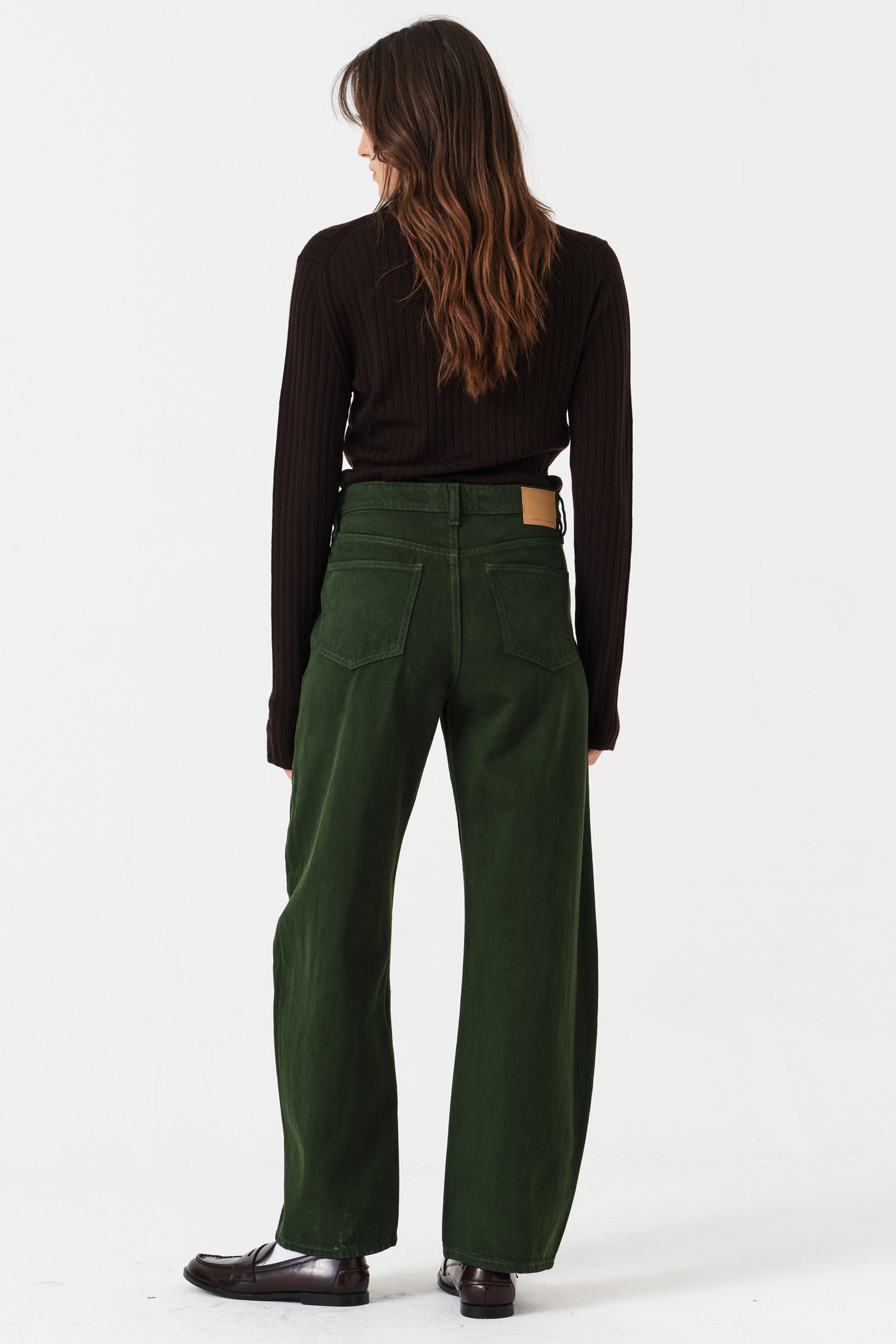 Woman wearing dark green barrel leg jeans and a brown shirt on a white background, photographed from the back