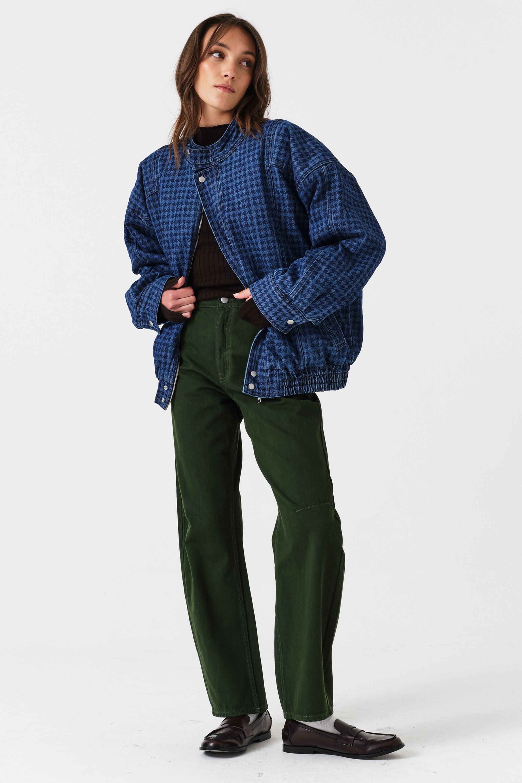 Woman wearing dark green barrel leg jeans, a brown shirt, and a dark blue houndstooth bomber jacket on a white background