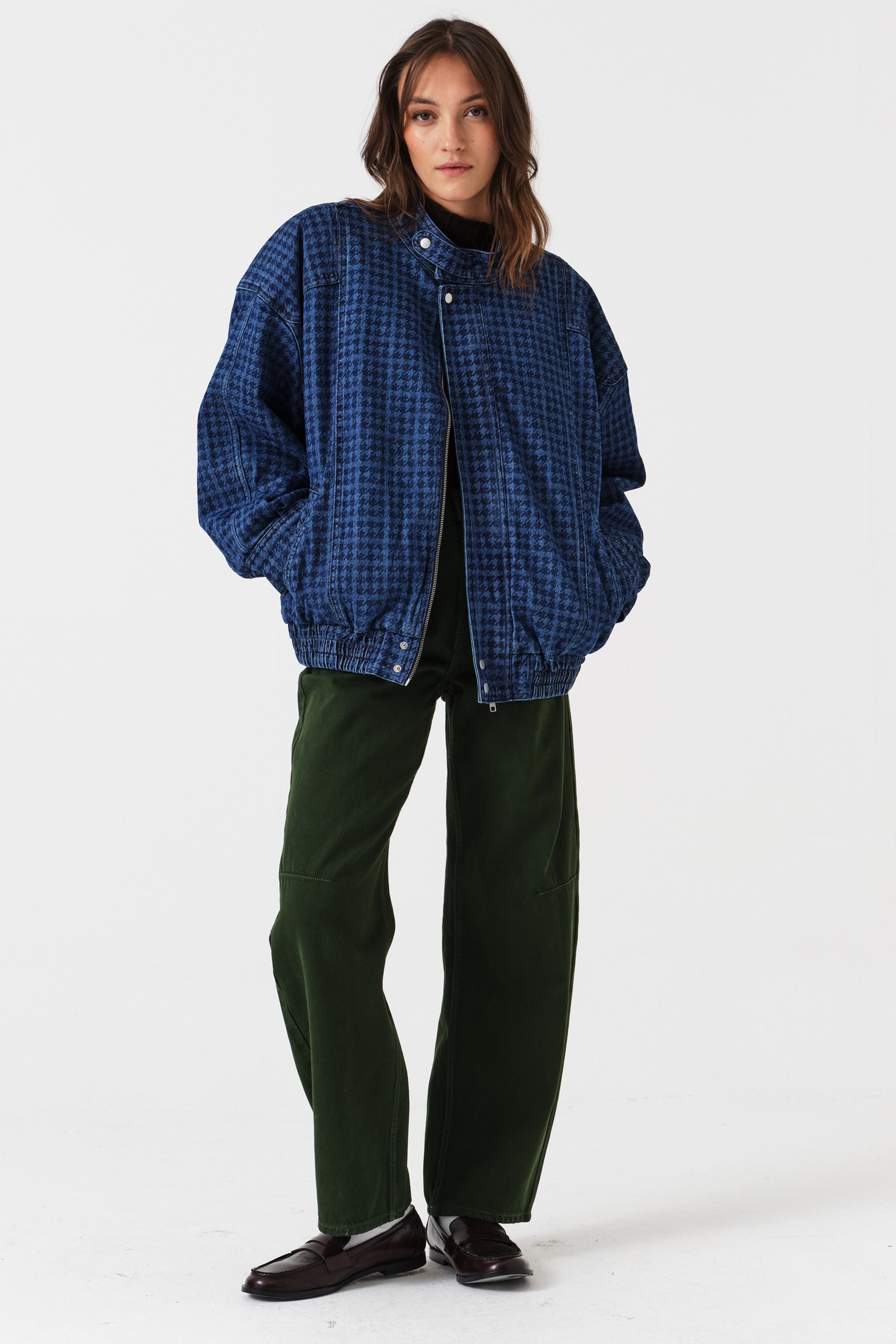 Woman wearing dark green barrel leg jeans, a brown shirt, and a dark blue houndstooth bomber jacket on a white background