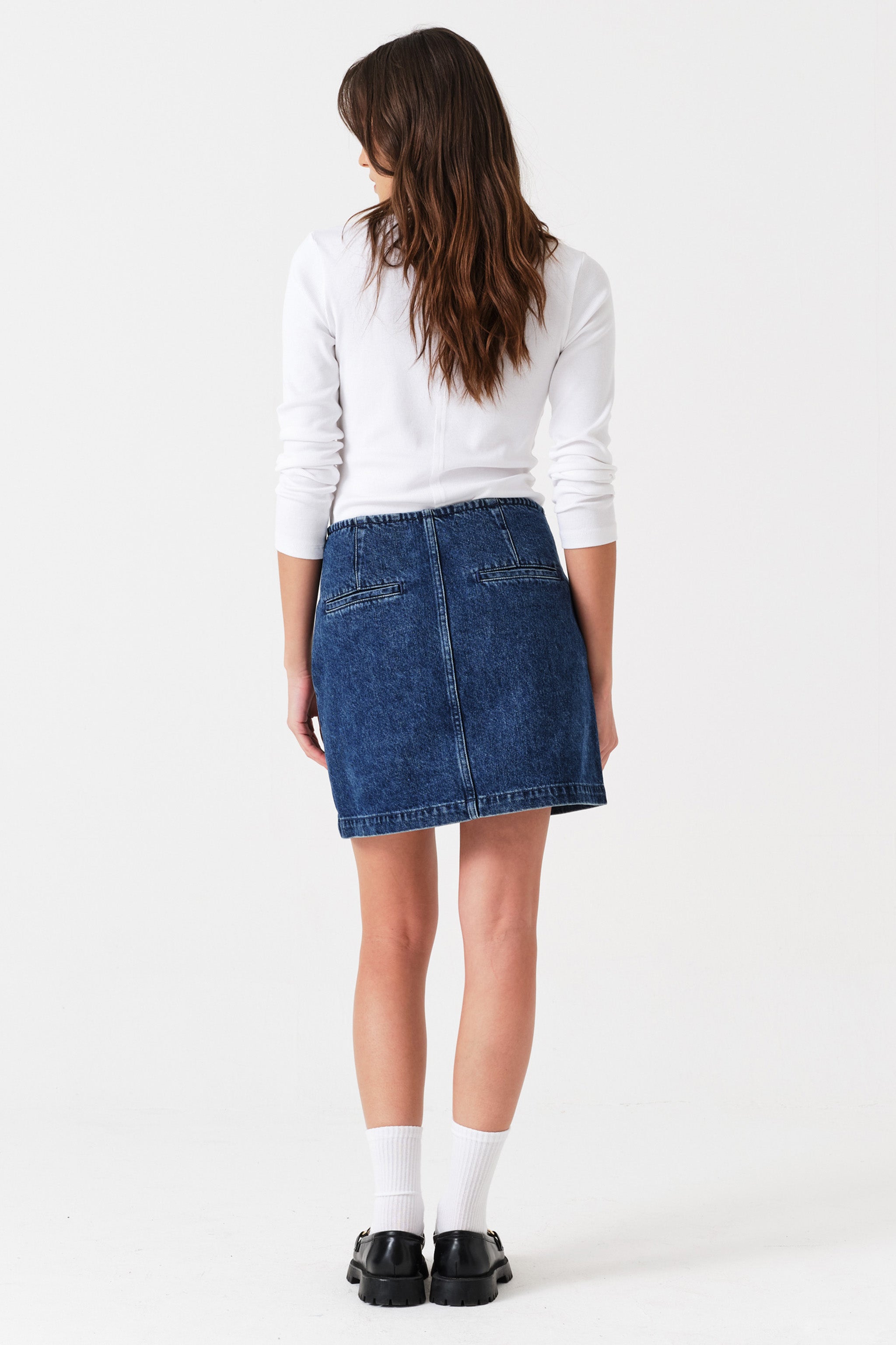 Woman wearing a dark blue denim mini skirt  and a white shirt on a white background, photographed from the back
