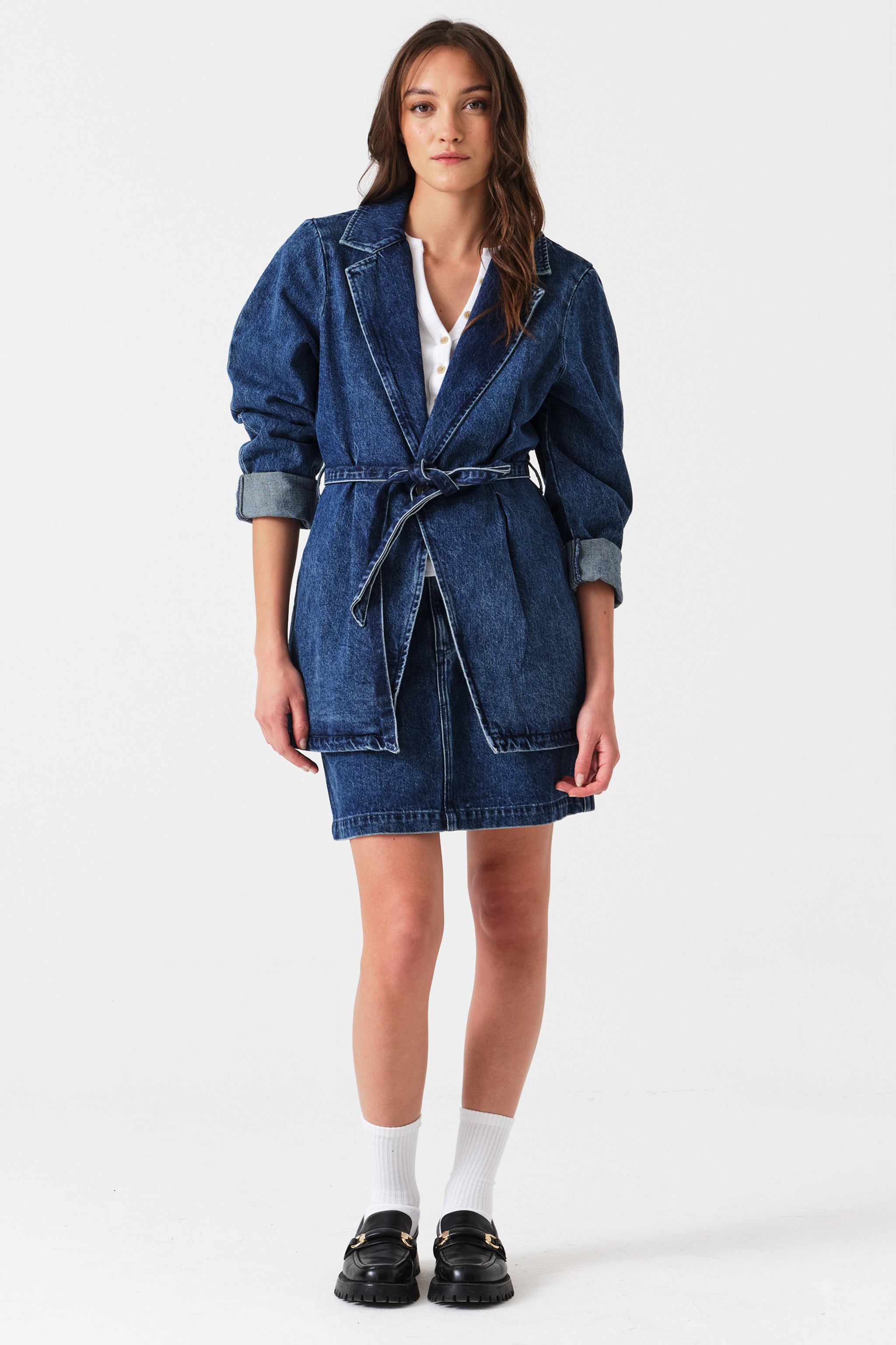 Woman wearing a dark blue denim mini skirt with seam details, a white shirt, and a dark blue denim blazer jacket with a tie waist on a white background