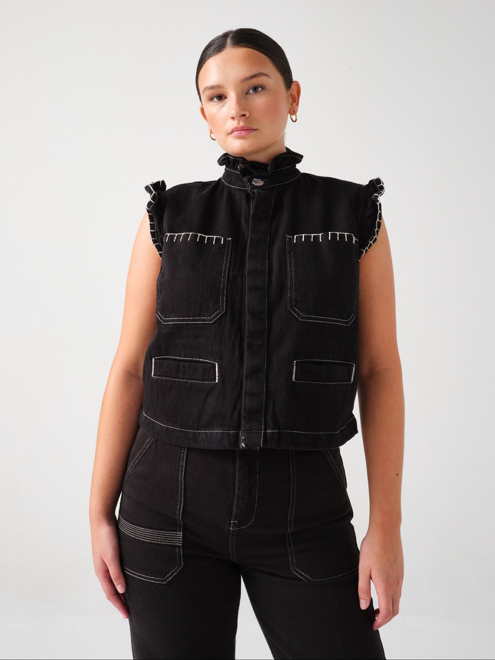 A woman wearing a black denim vest with a frill collar, white blanket stitch details, and two large patch pockets, paired with black denim trousers against a white background