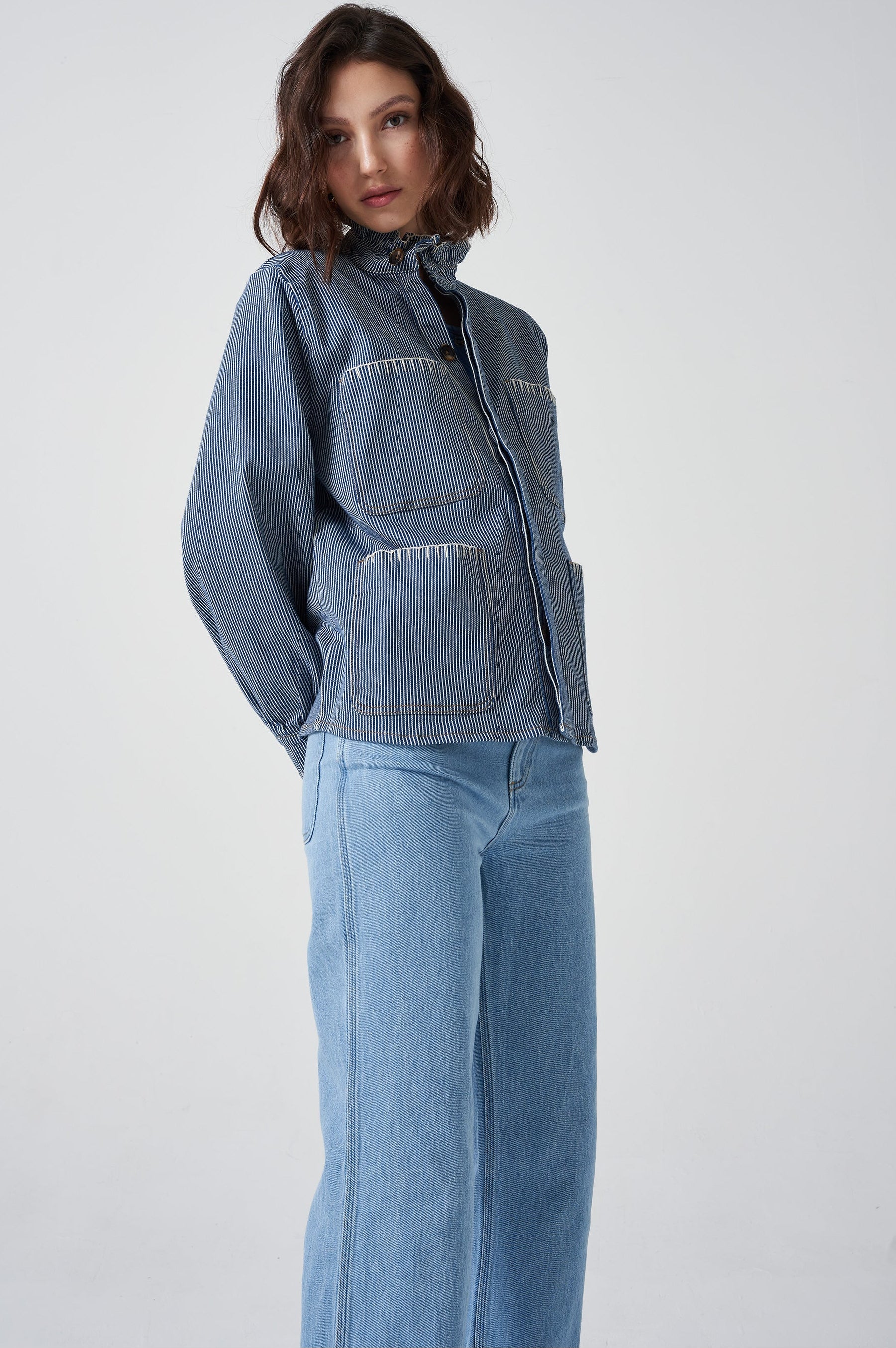 A woman wearing a blue and white striped jacket with a frill collar and our signature white blanket stitching on four large patch pockets, paired with blue jeans against a white background