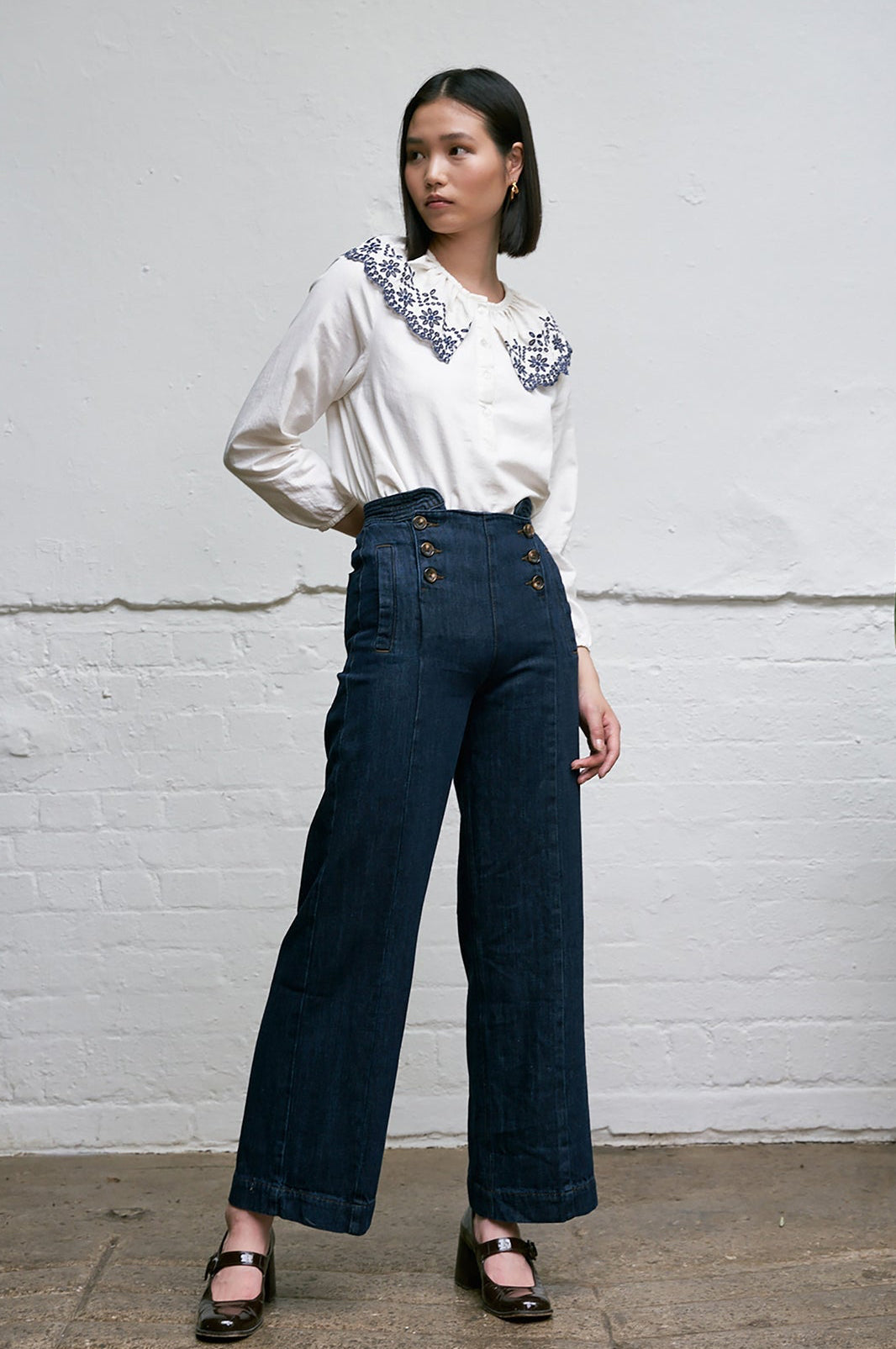 Woman wearing a white blouse with blue floral embroidery details on an eyelet style collar with blue jeans against a white background