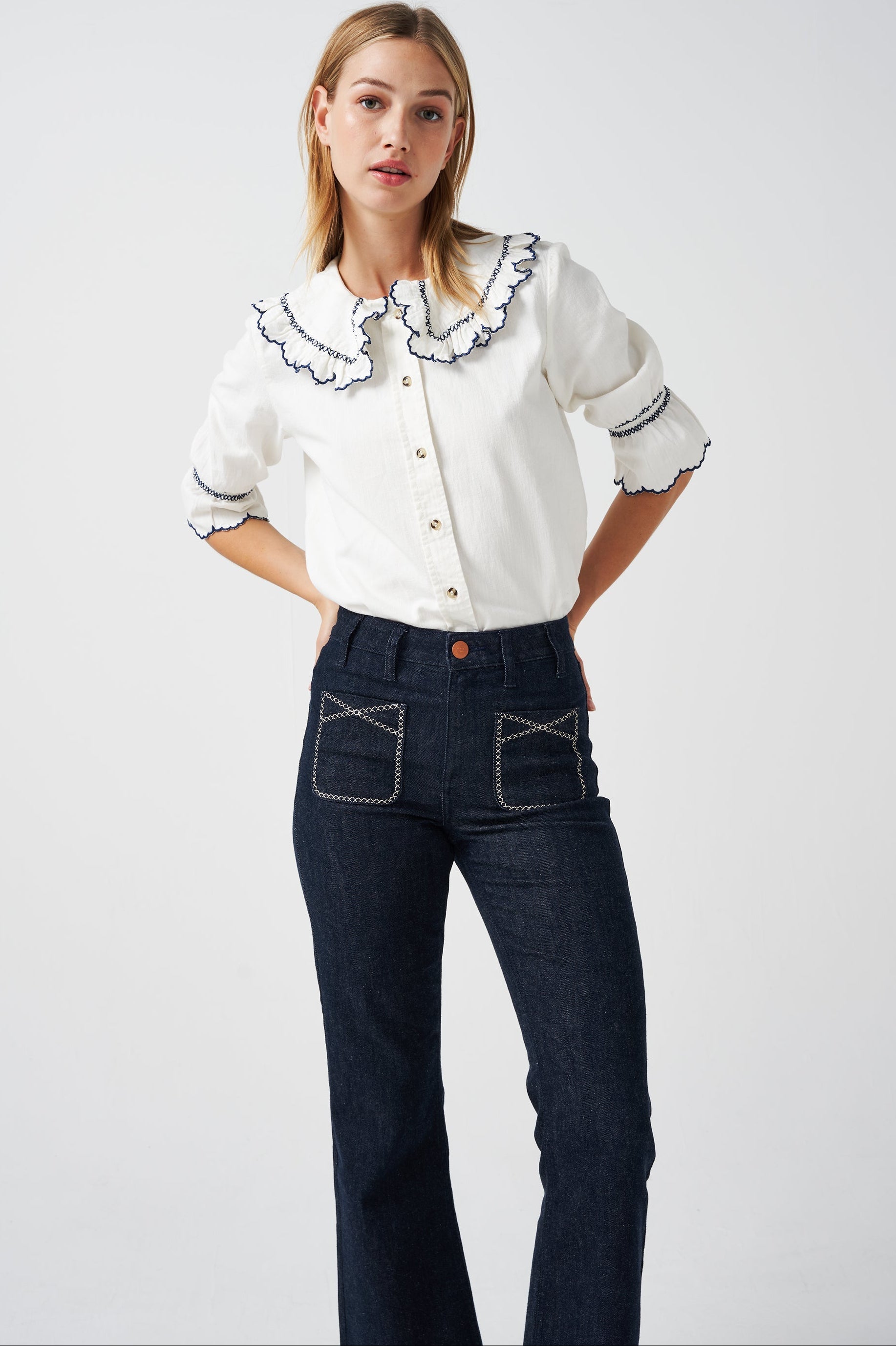 A woman wearing a white organic cotton blouse with dark blue embroiderery on frilled sleeves and a frill peek-a-boo collar with dark blue jeans on a white background