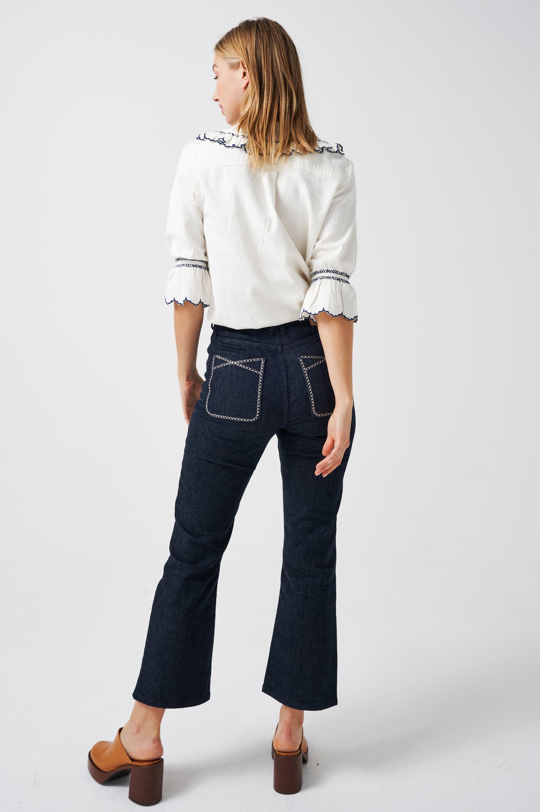 A woman wearing a white organic cotton blouse with dark blue embroiderery on frilled sleeves and a frill peek-a-boo collar with dark blue jeans on a white background