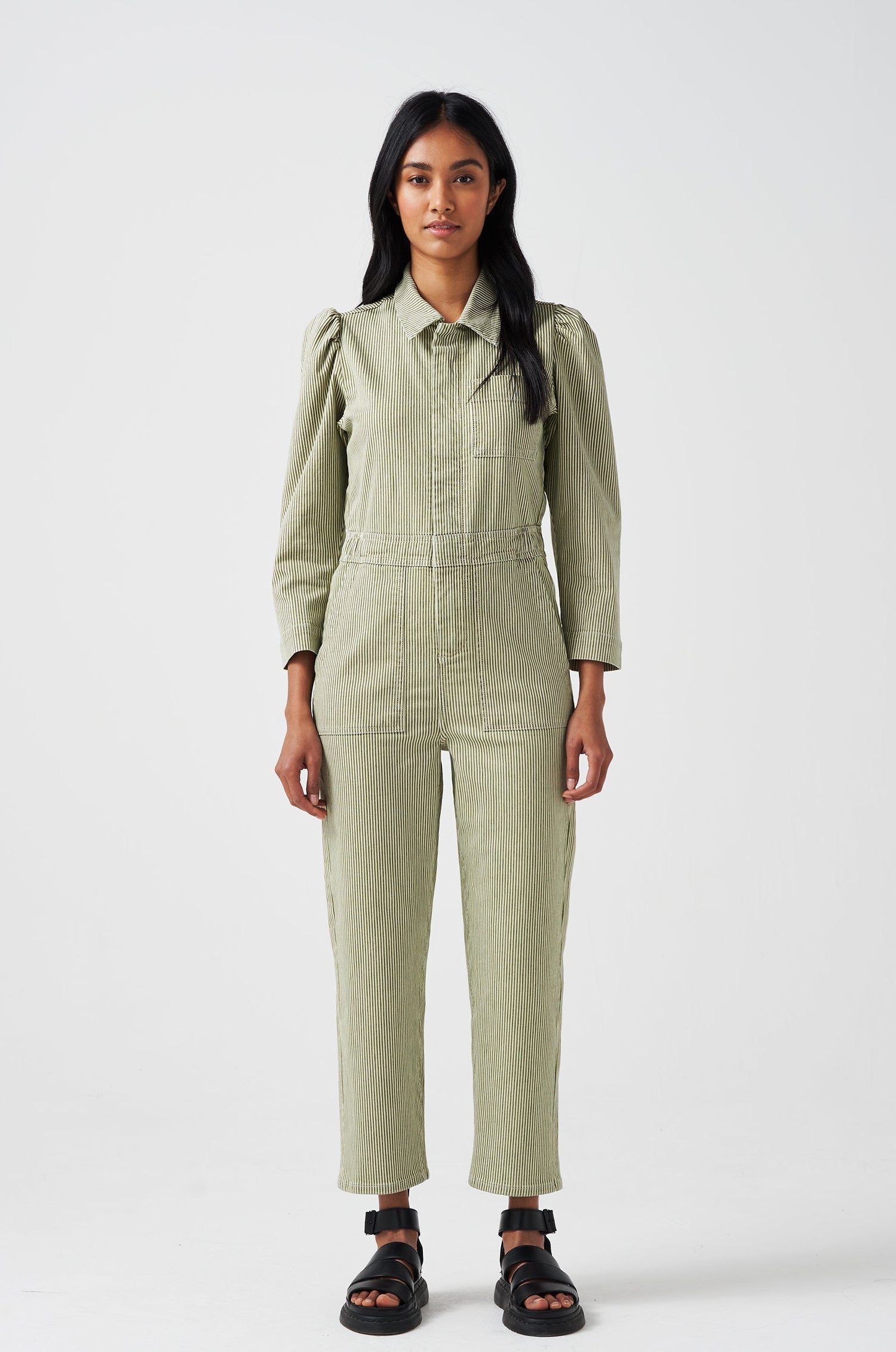 A woman wearing a khaki green and white striped jumpsuit with soft puff shoulders, large pockets, and a front zip detail on a white background