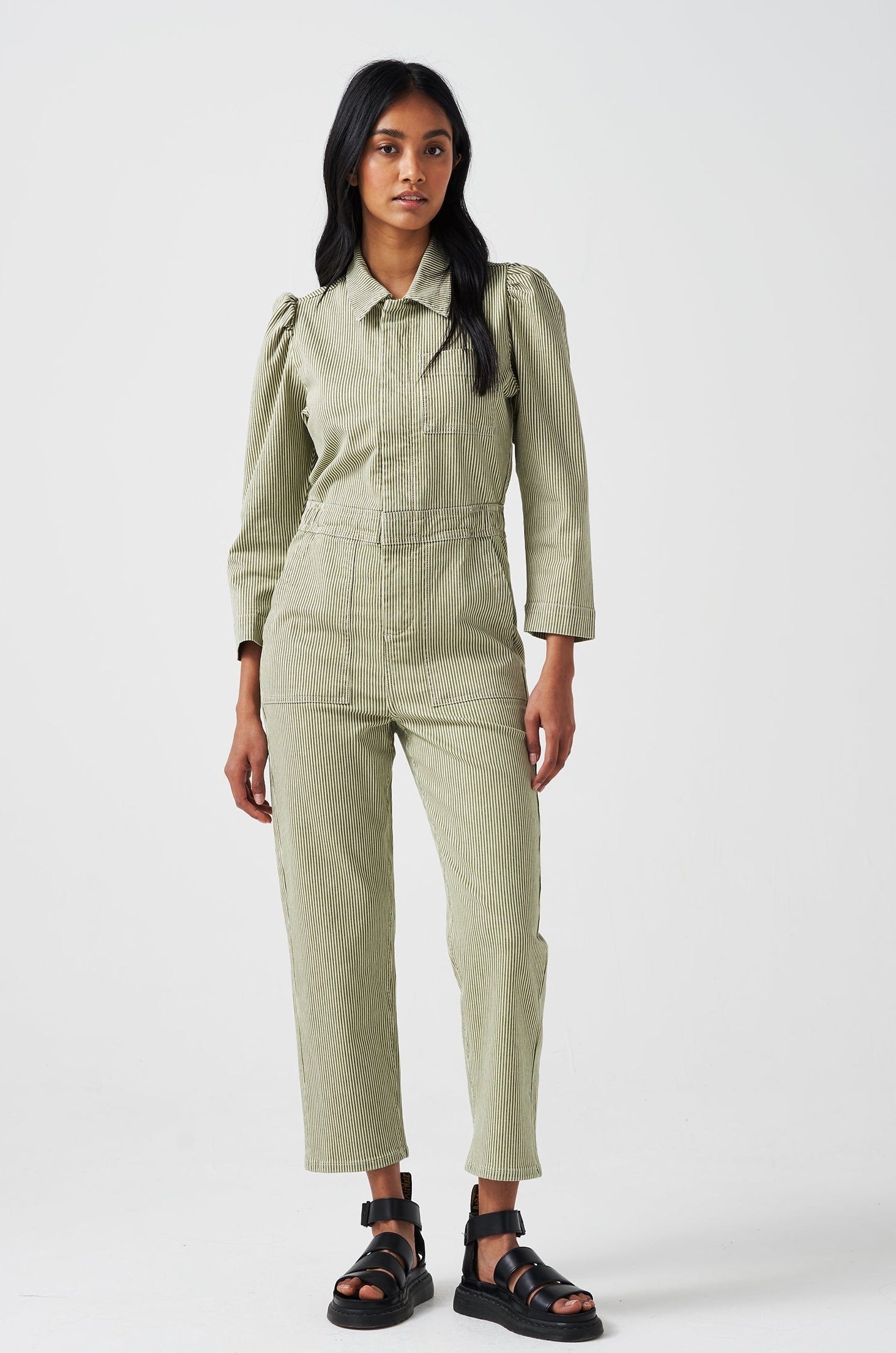 A woman wearing a khaki green and white striped jumpsuit with soft puff shoulders, large pockets, and a front zip detail on a white background