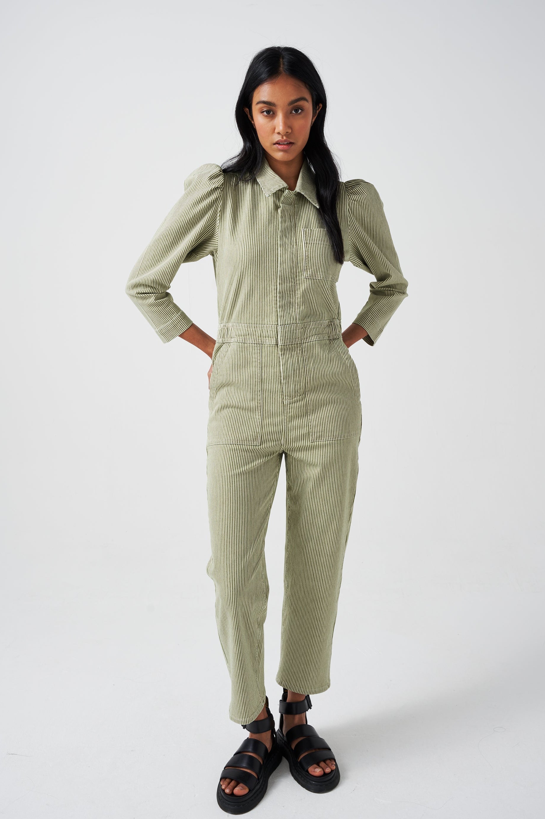 A woman wearing a khaki green and white striped jumpsuit with soft puff shoulders, large pockets, and a front zip detail on a white background