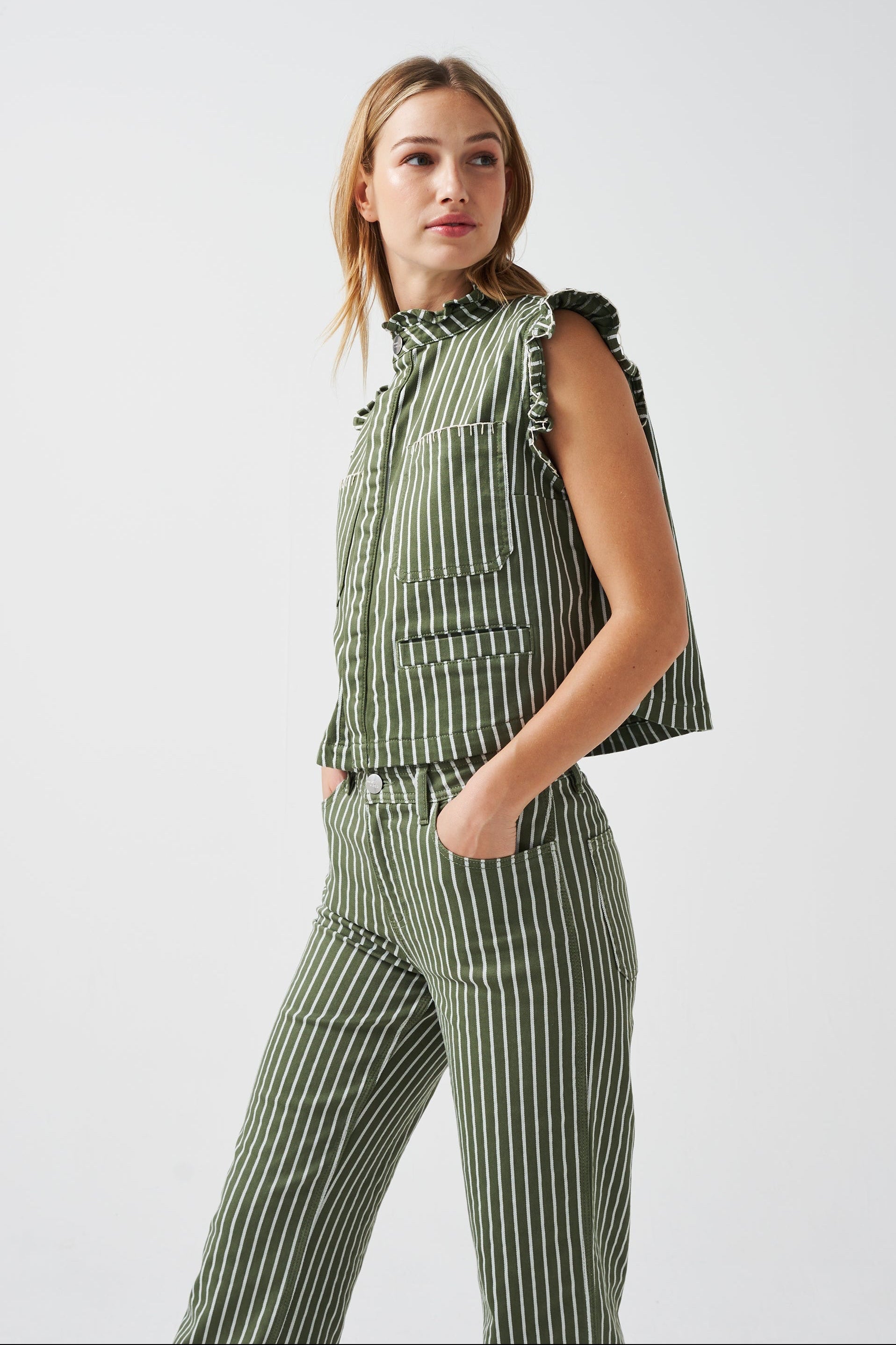 A woman wearing a khaki and white striped vest/ waistcoat with a frill collar, white blanket stitching, and two large patch pockets, part of a matching set, on a white background.