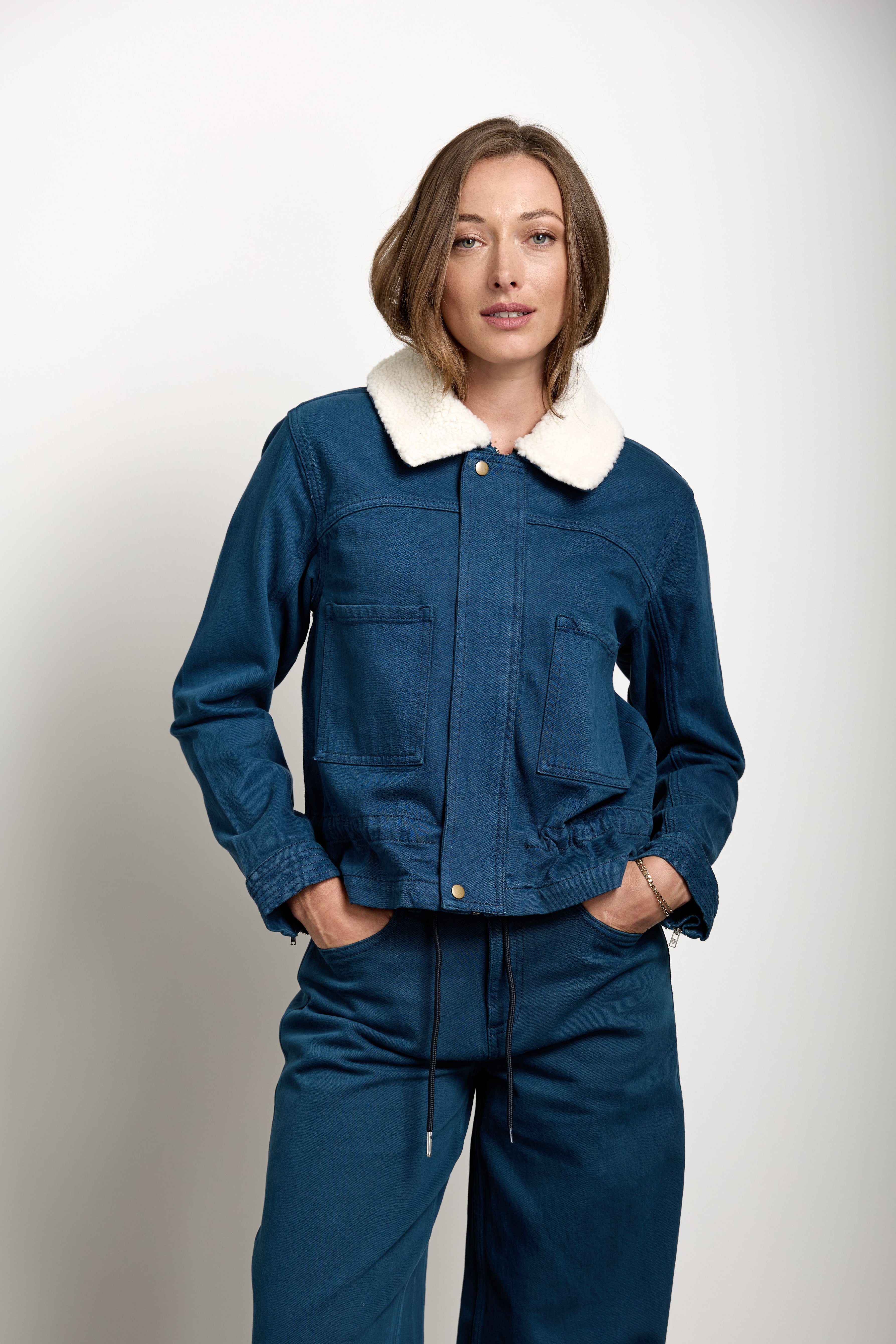 A woman modeling a dark blue denim jacket with a contrasting cream-colored fleece collar, paired with matching dark blue denim pants, against a plain light gray studio background.