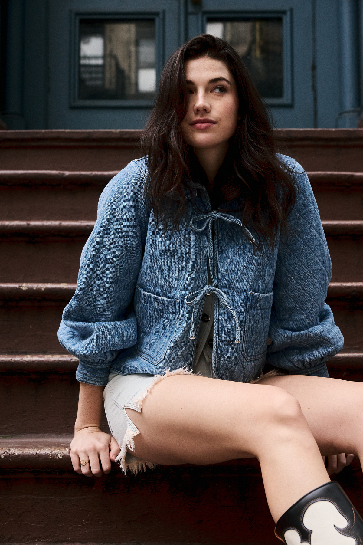 A woman wearing a light blue quilted denim jacket with a frill collar, front tie closures, and two large pockets paired with a white shirt and white shorts on doorway steps