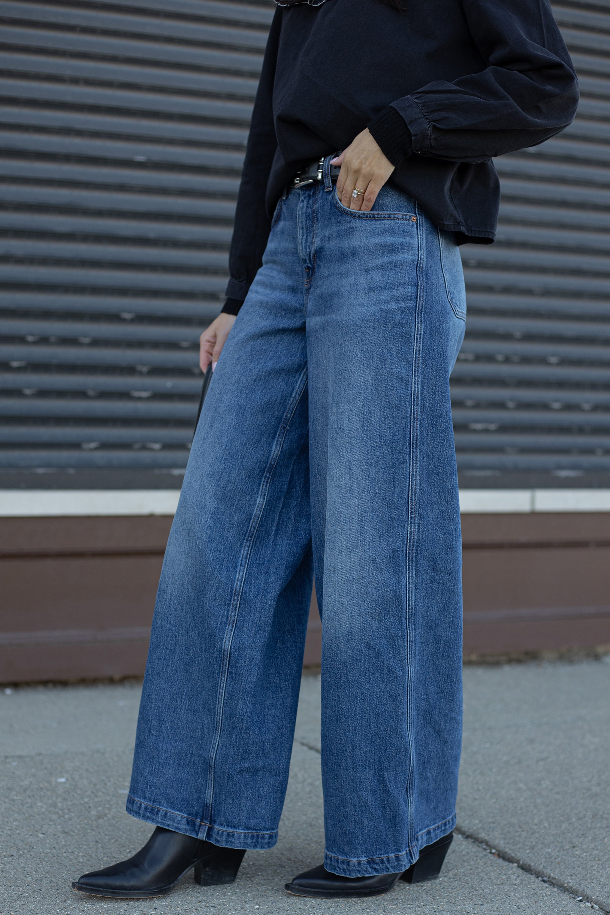 woman wearing blue wide-leg jeans and a black top against a metallic wall.