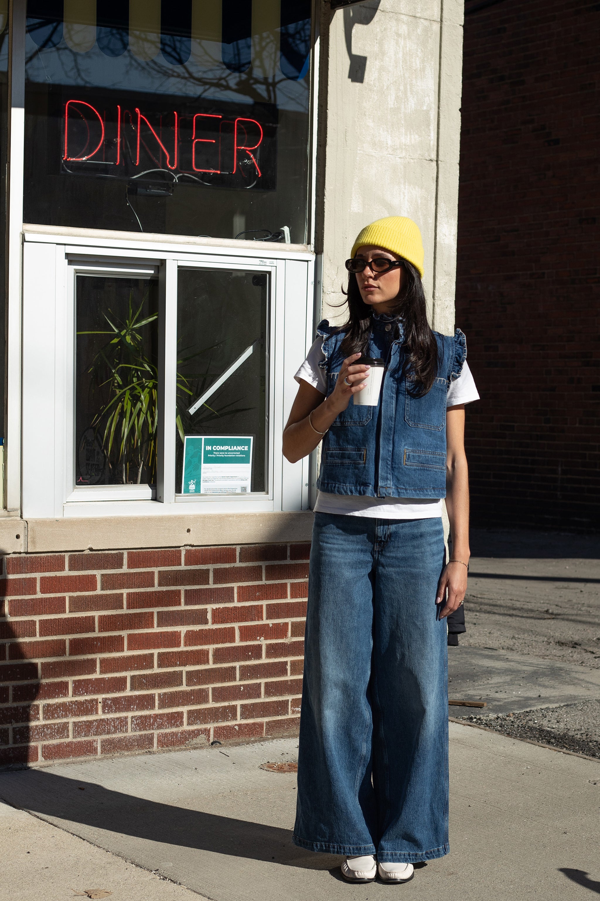 Woman holding a coffee cup and wearing a denim vest with frill details, white shirt, and blue wide leg jeans against a diner sign.