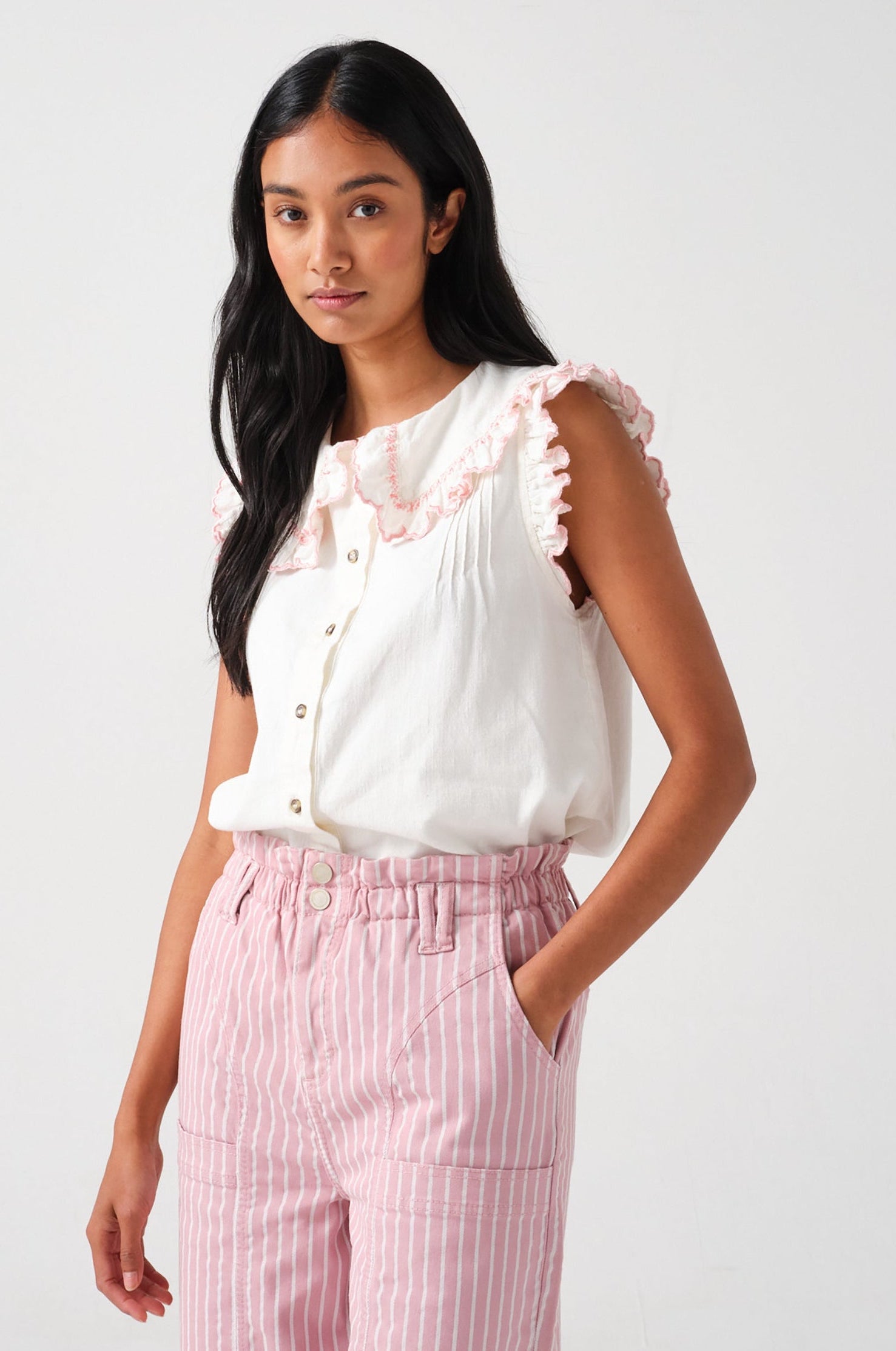 A woman wearing a short sleeve blouse with a frill collar and embroidered details in ecru and dusty rose colors, paired with striped pants.